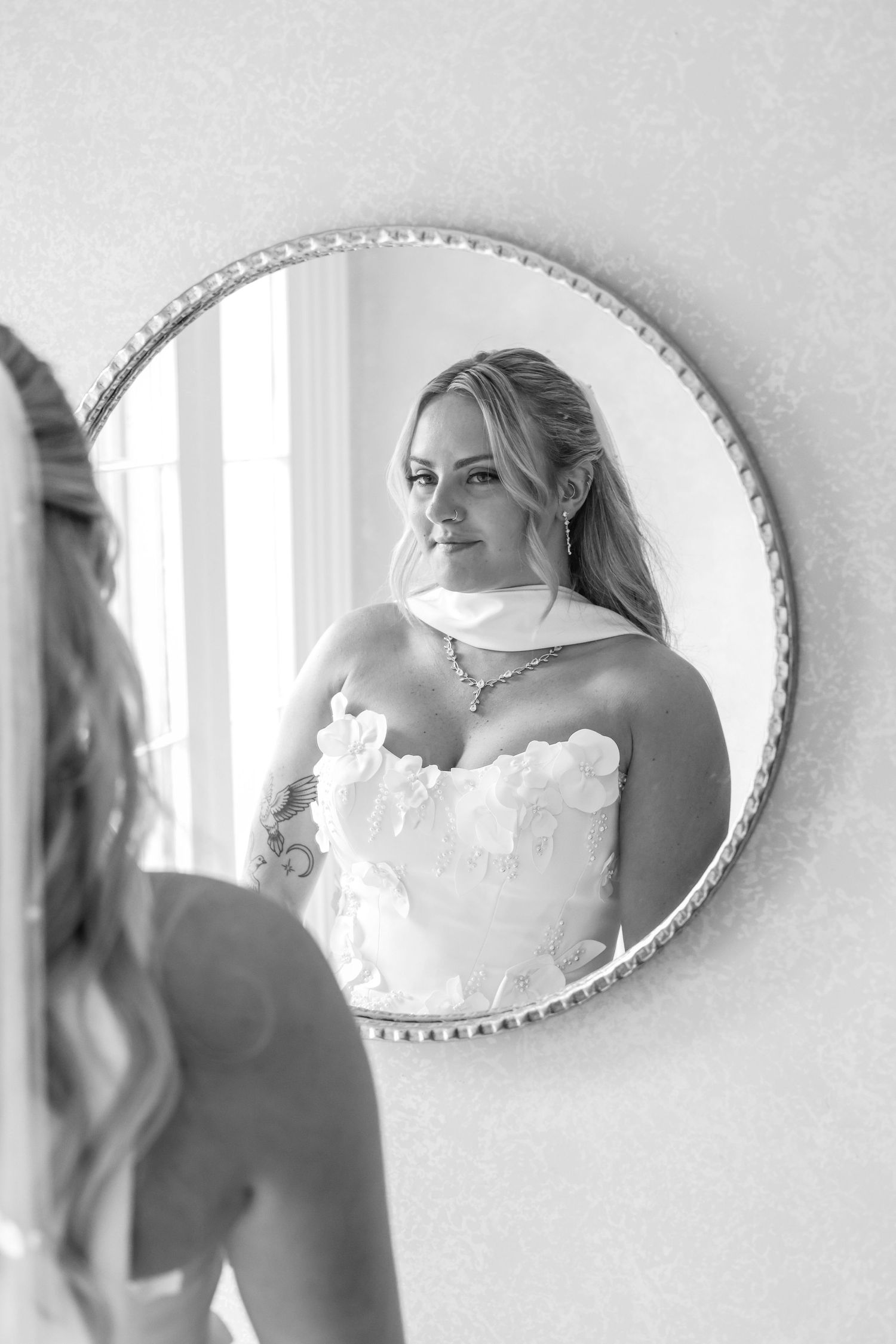 Black and white photo of a bride in a strapless gown admiring her reflection in a round mirror.