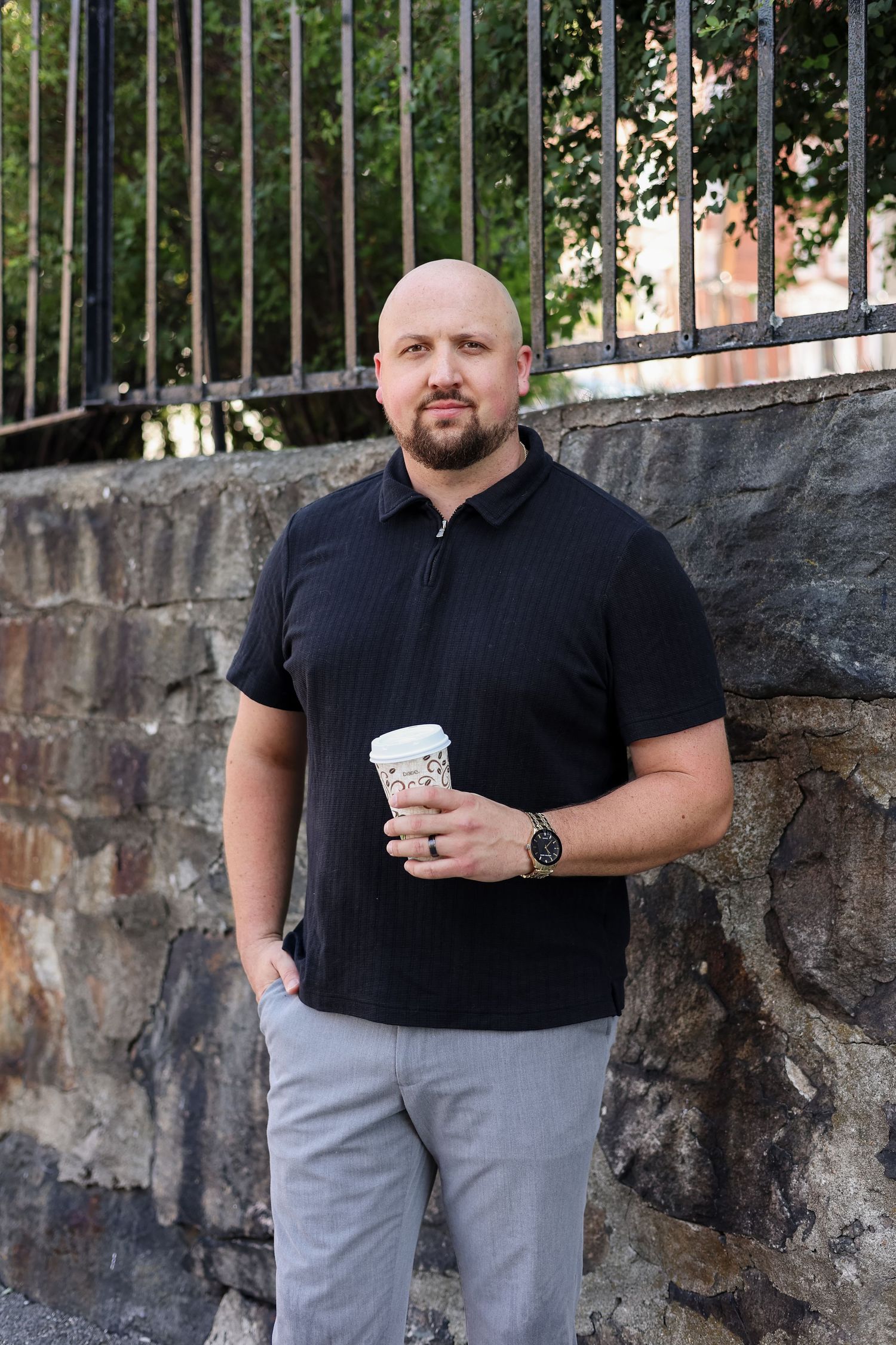Bald man with beard holding coffee cup, wearing black polo and gray pants, standing against stone wall outdoors.