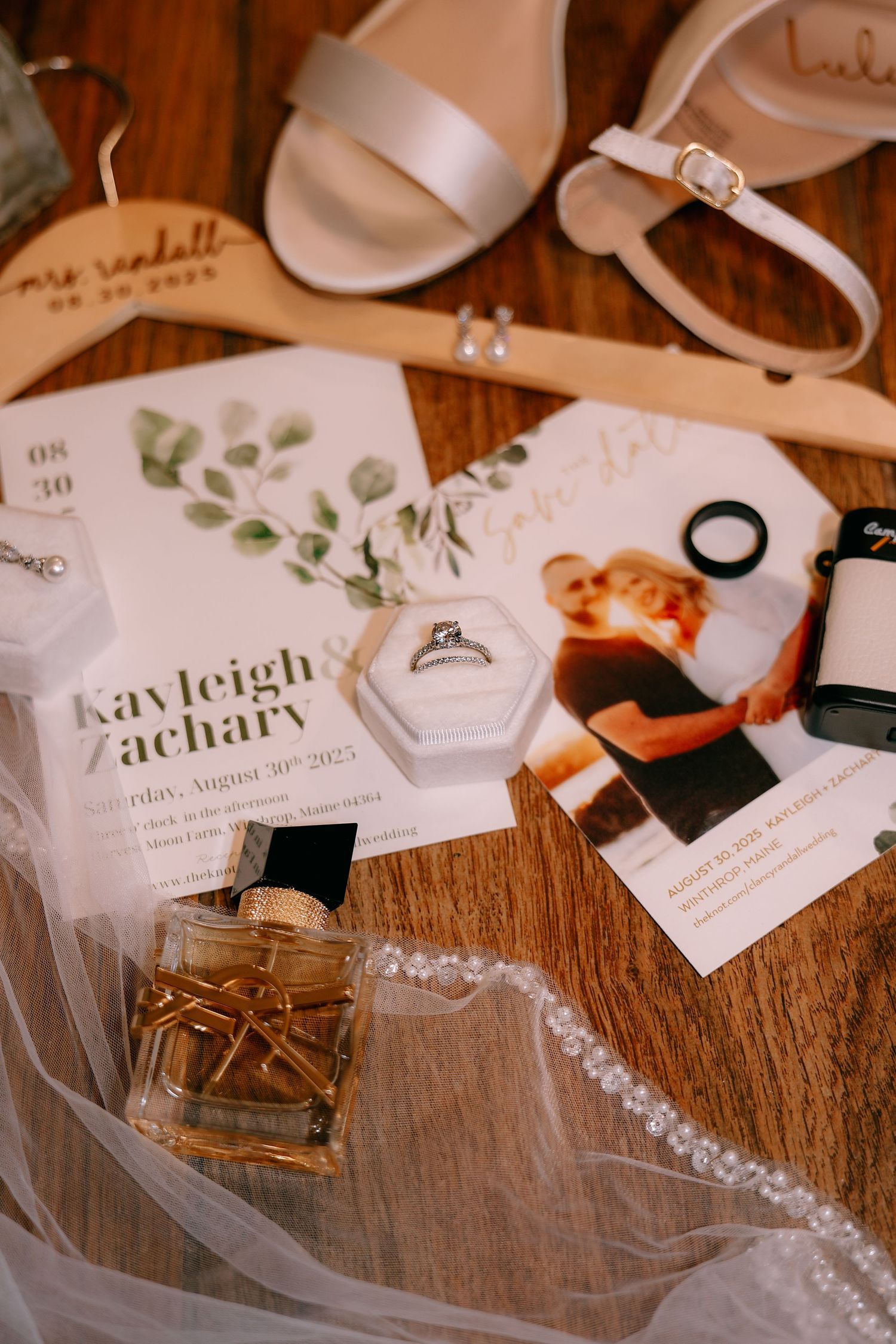 Wedding details flatlay with invitation, shoes, perfume and accessories on wooden surface.