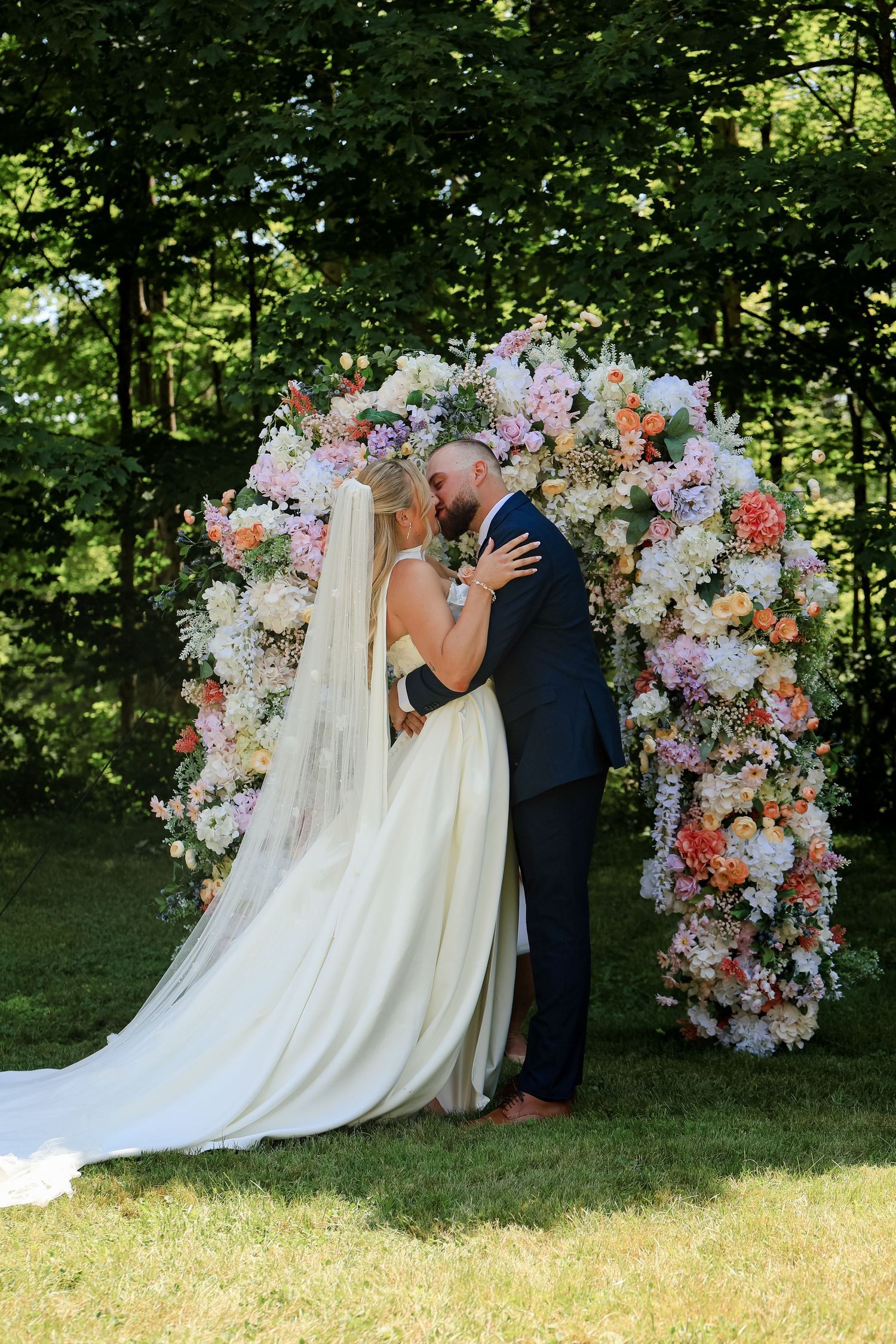 Bride and groom share first kiss under a colorful floral arch outdoors, surrounded by lush greenery on their wedding day.