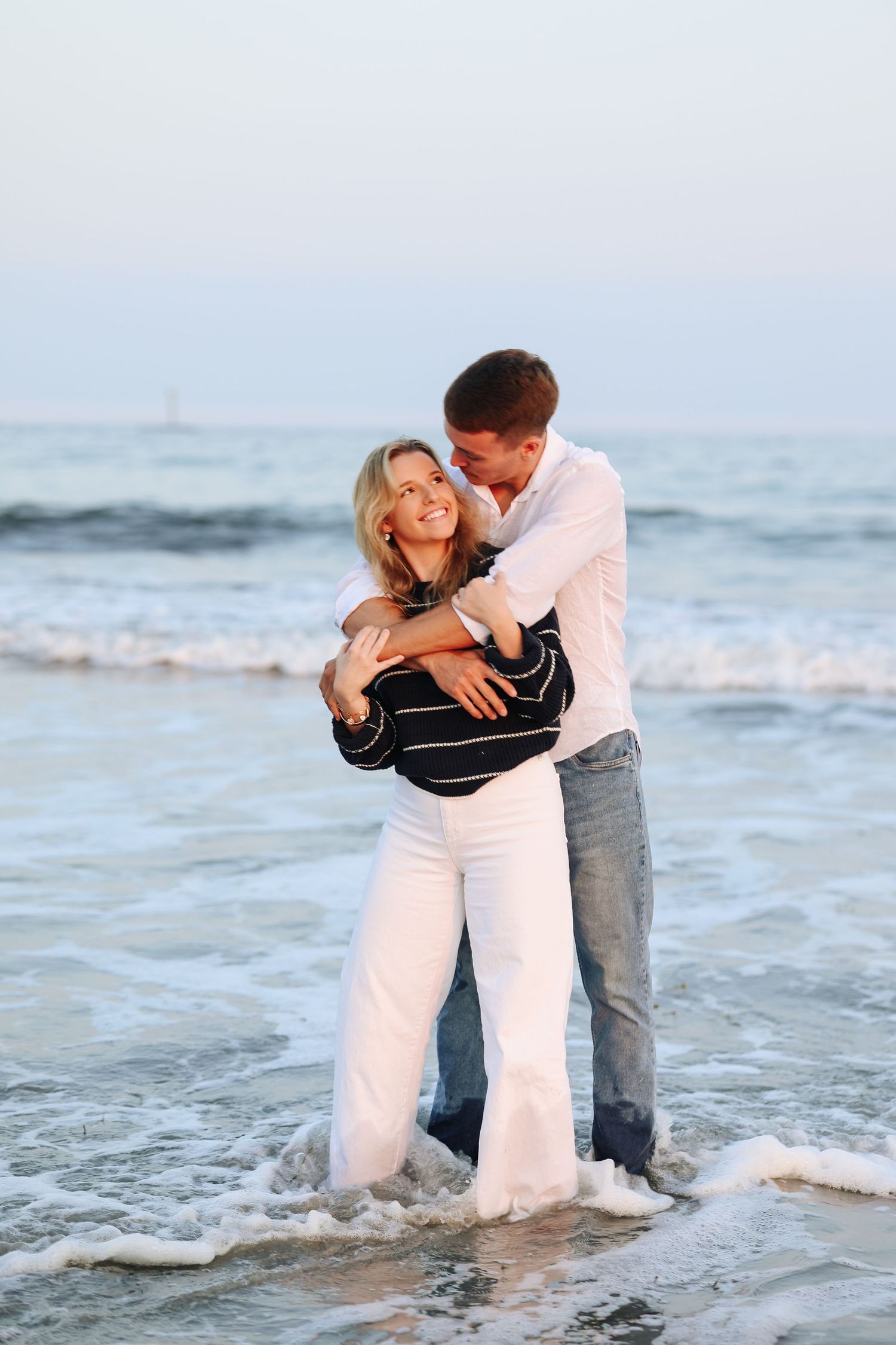 Couple embracing and laughing together while standing in ocean waves on a beach at sunset.