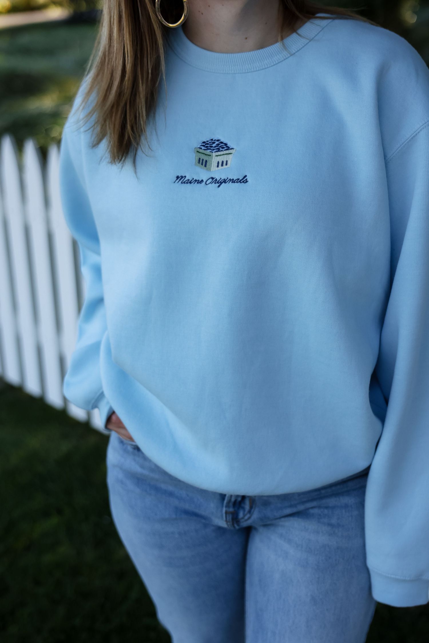 Person wearing a light blue crewneck sweatshirt with embroidered Maine Originals logo, paired with blue jeans, standing by a white picket fe