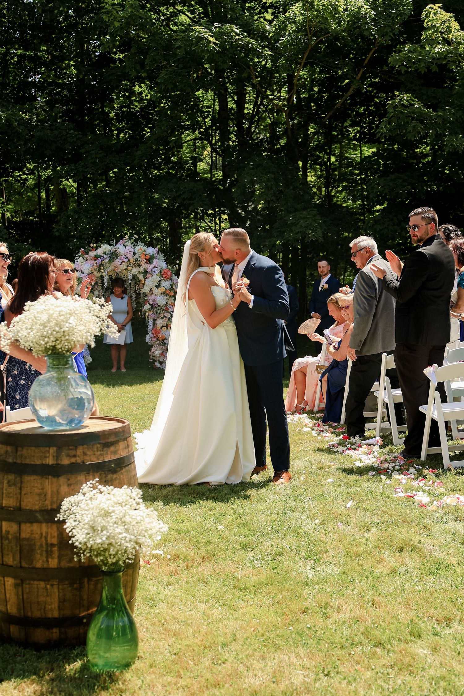 Bride and groom share first kiss at outdoor wedding ceremony with floral arch, guests standing, and rustic barrel with flowers.