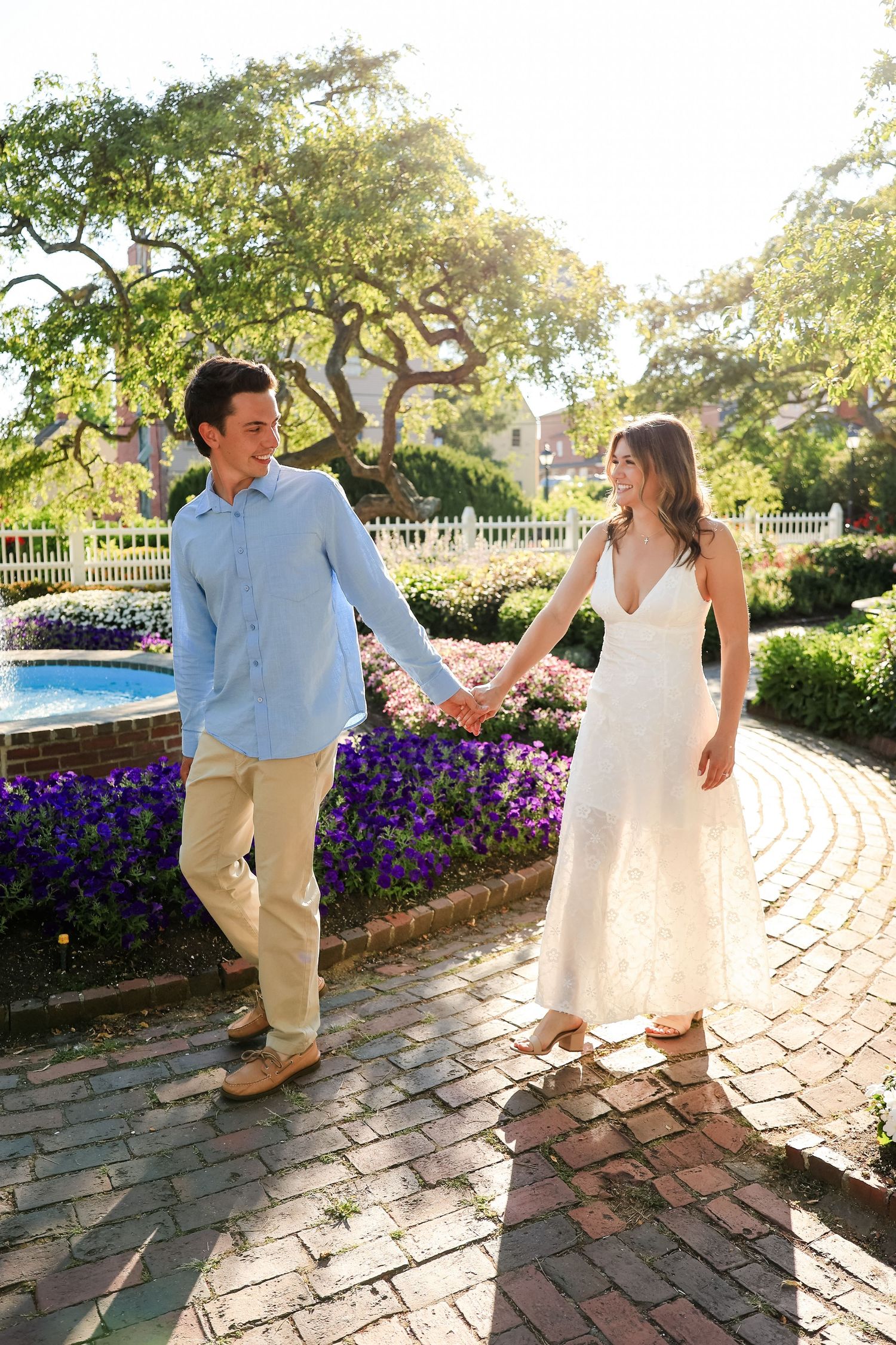 Couple holding hands walking on brick path in garden with purple flowers and fountain backdrop.