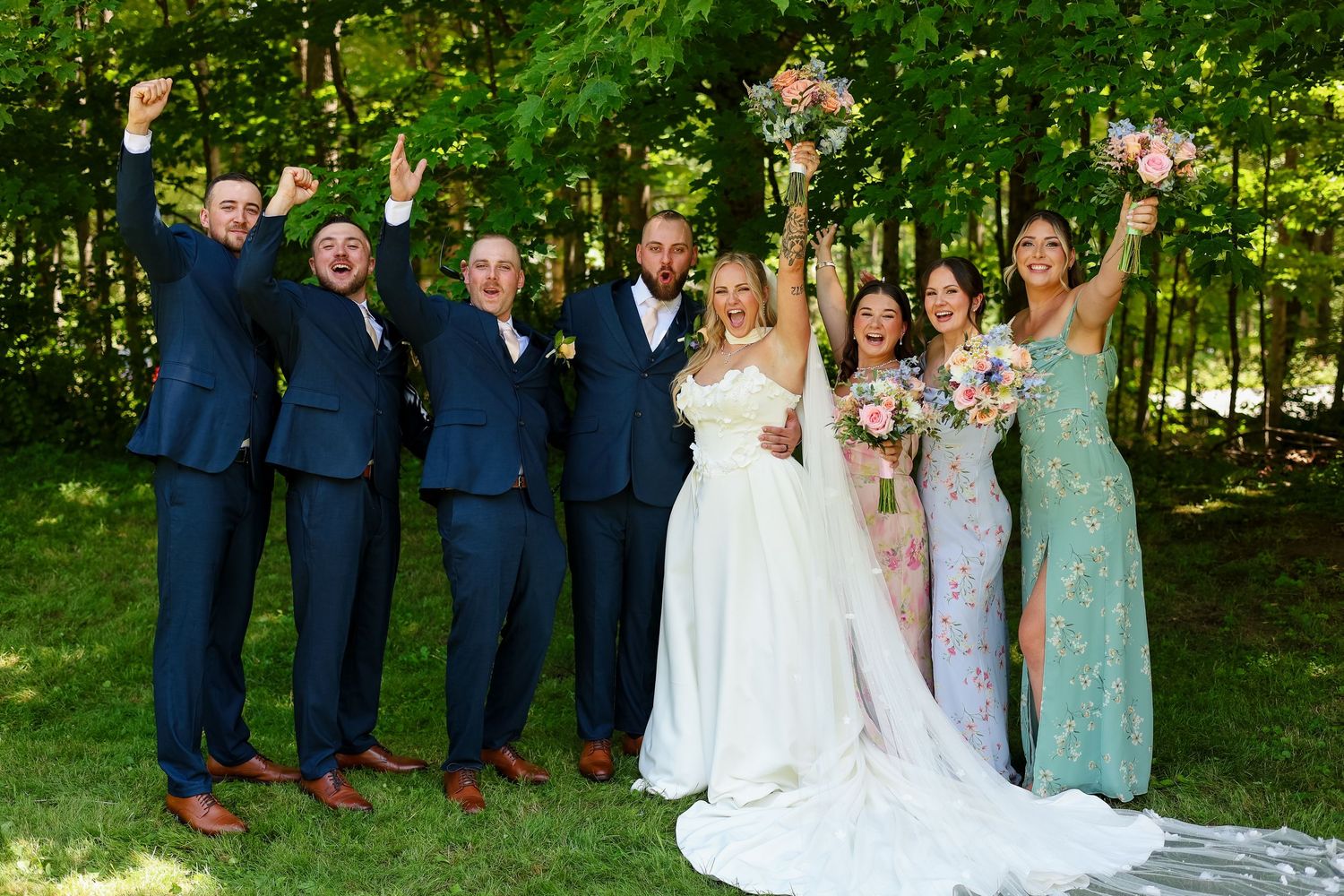 Wedding party celebrating outdoors, bride and groom with groomsmen in navy suits and bridesmaids in sage dresses, arms raised.