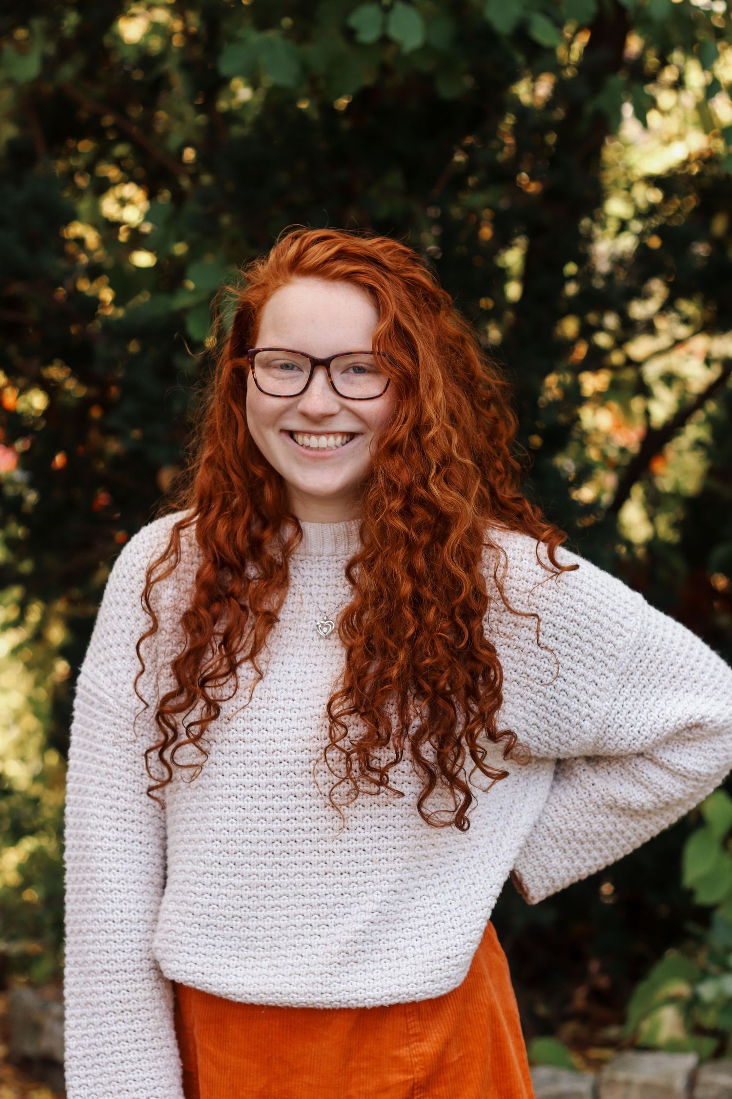 Solo female portrait in a cream sweater with curly red hair poses outdoors against a natural background.
