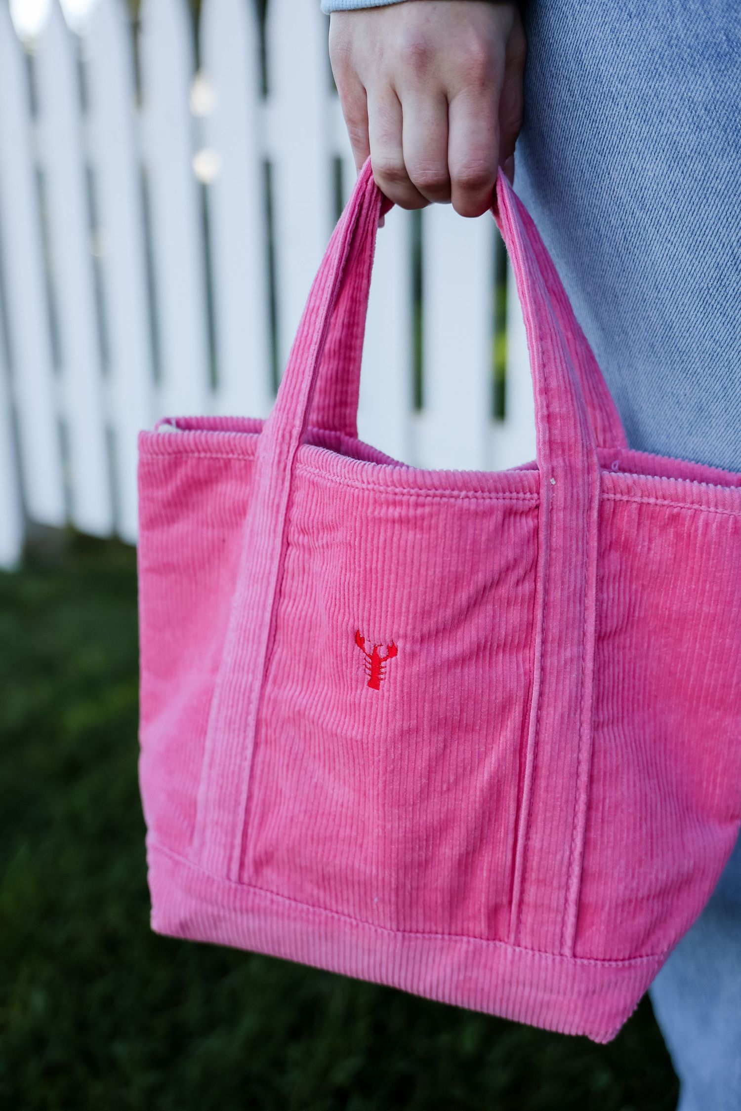 Hand holding a pink corduroy mini tote bag with a small red embroidered lobster, in front of a white picket fence.
