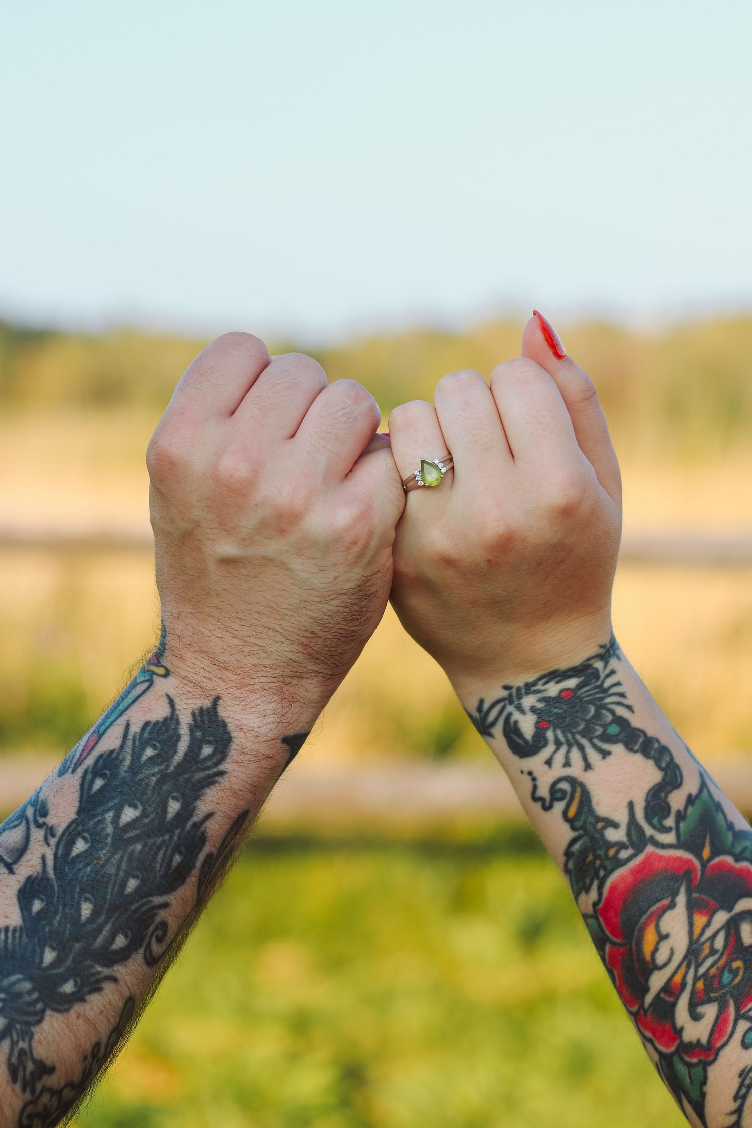 Two tattooed hands interlock pinkies outdoors, one wearing a gold ring with red nails, against a blurred natural background.