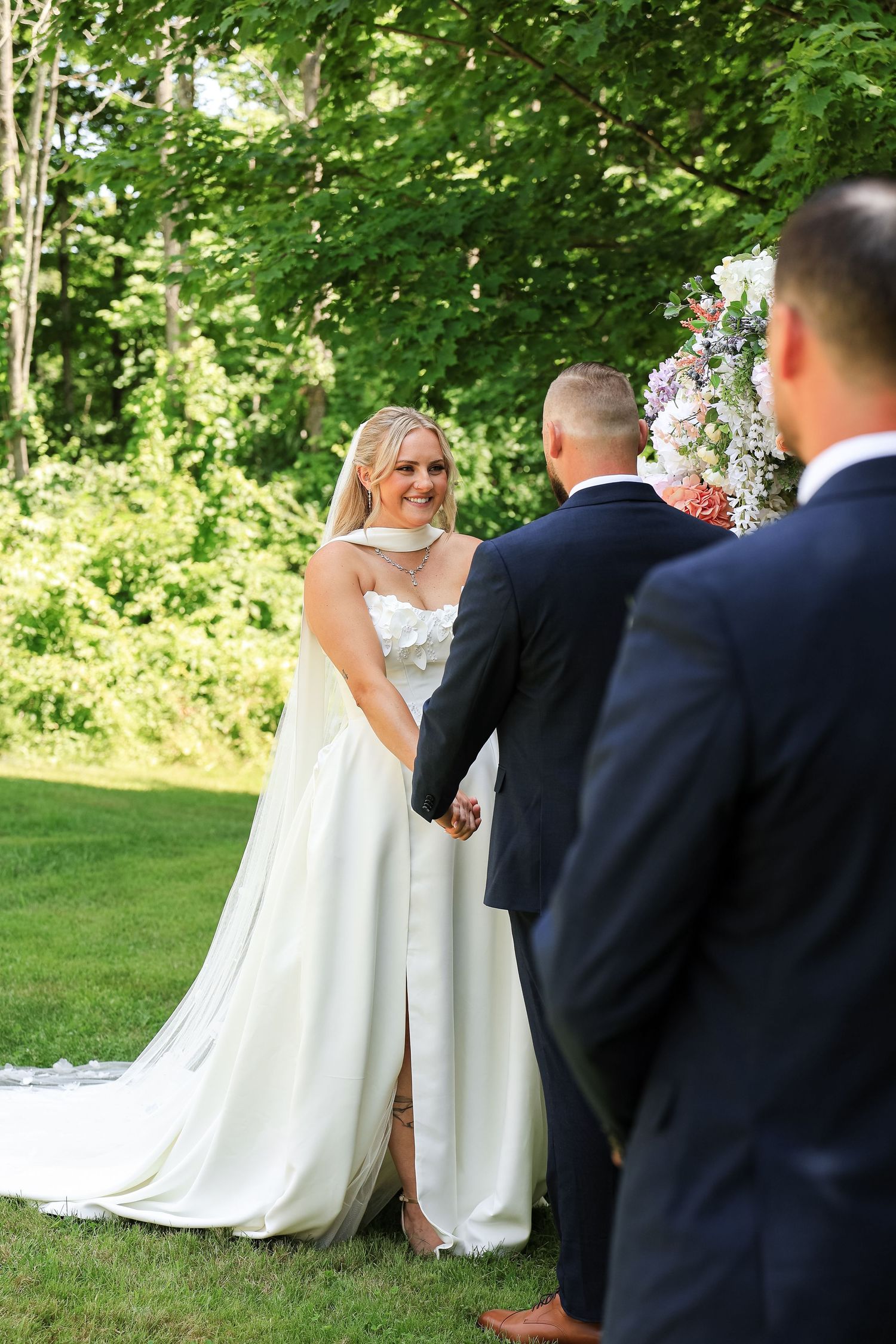 Bride in white gown smiles at groom during outdoor wedding ceremony surrounded by lush green trees.