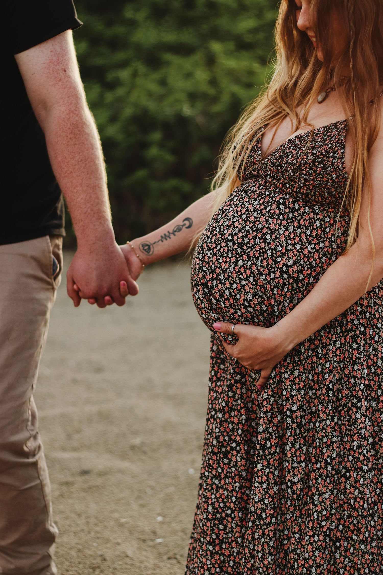 Pregnant woman in floral dress holds hands with partner outdoors, cradling baby bump during maternity photoshoot.