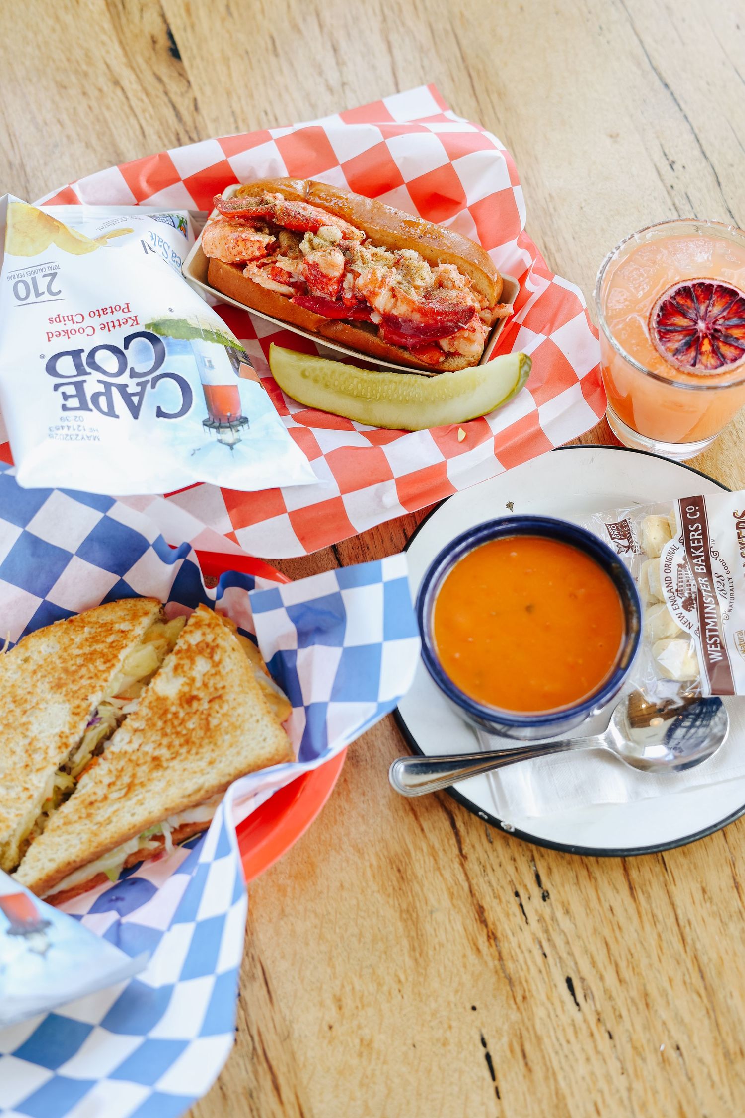 New England lobster roll, grilled sandwich, tomato soup, Cape Cod chips, and a cocktail served on a wooden table.
