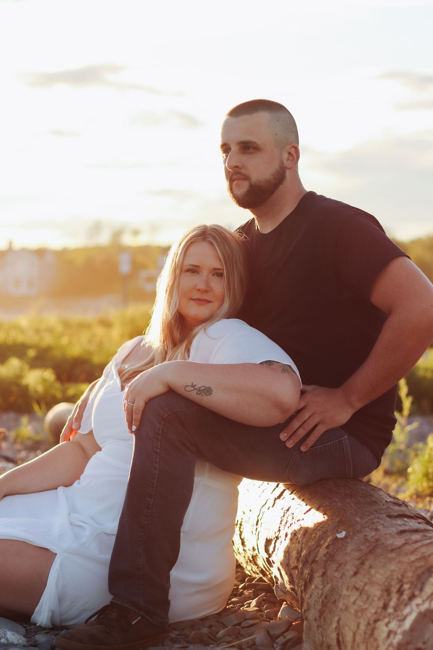 Couple sitting on a log at sunset, woman in white dress leaning against man in black shirt, golden hour outdoor portrait.