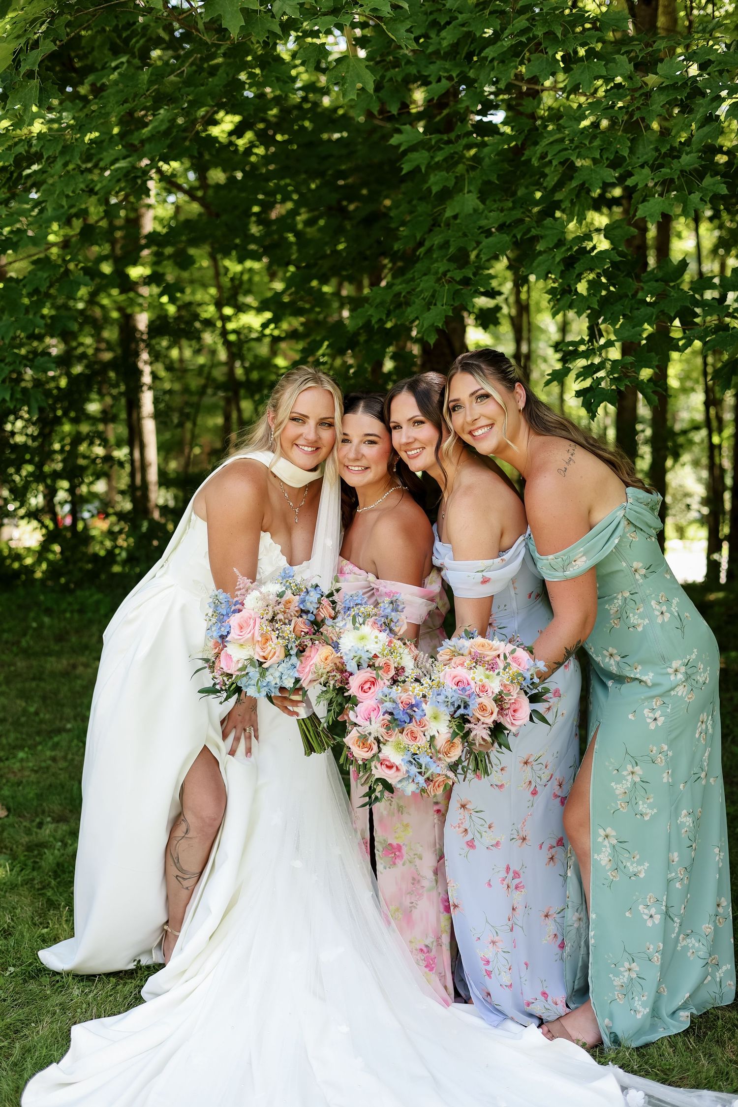 Bride in white gown poses with three bridesmaids in colorful floral dresses holding bouquets outdoors.