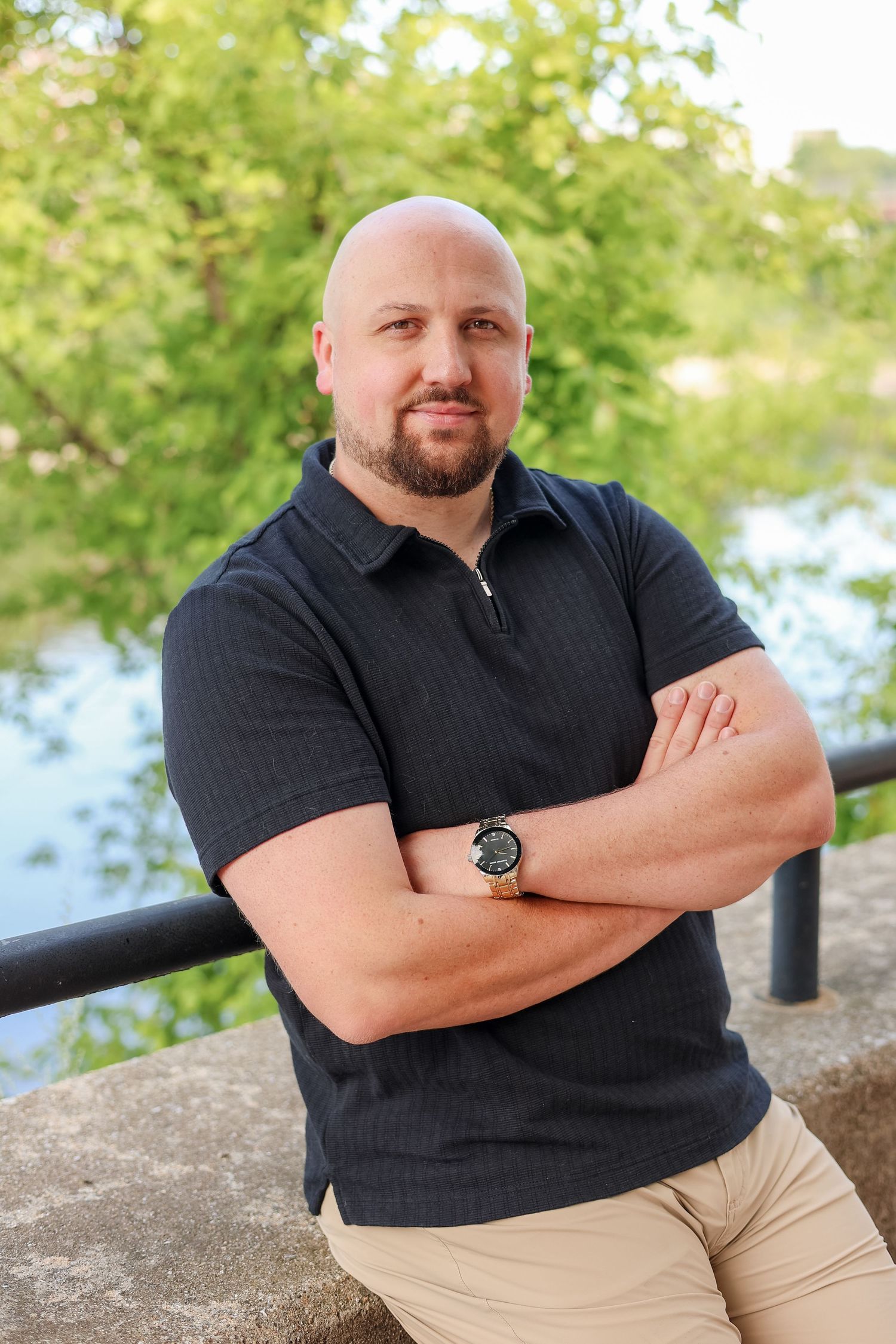 Bald man with beard, arms crossed, wearing dark polo and watch, posing outdoors near a railing with greenery.