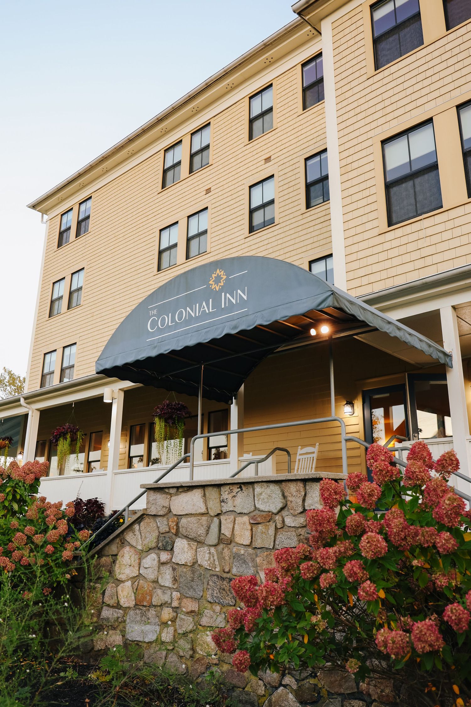Colonial Inn building exterior with beige siding, dark blue awning, and pink hydrangeas blooming by stone foundation steps.