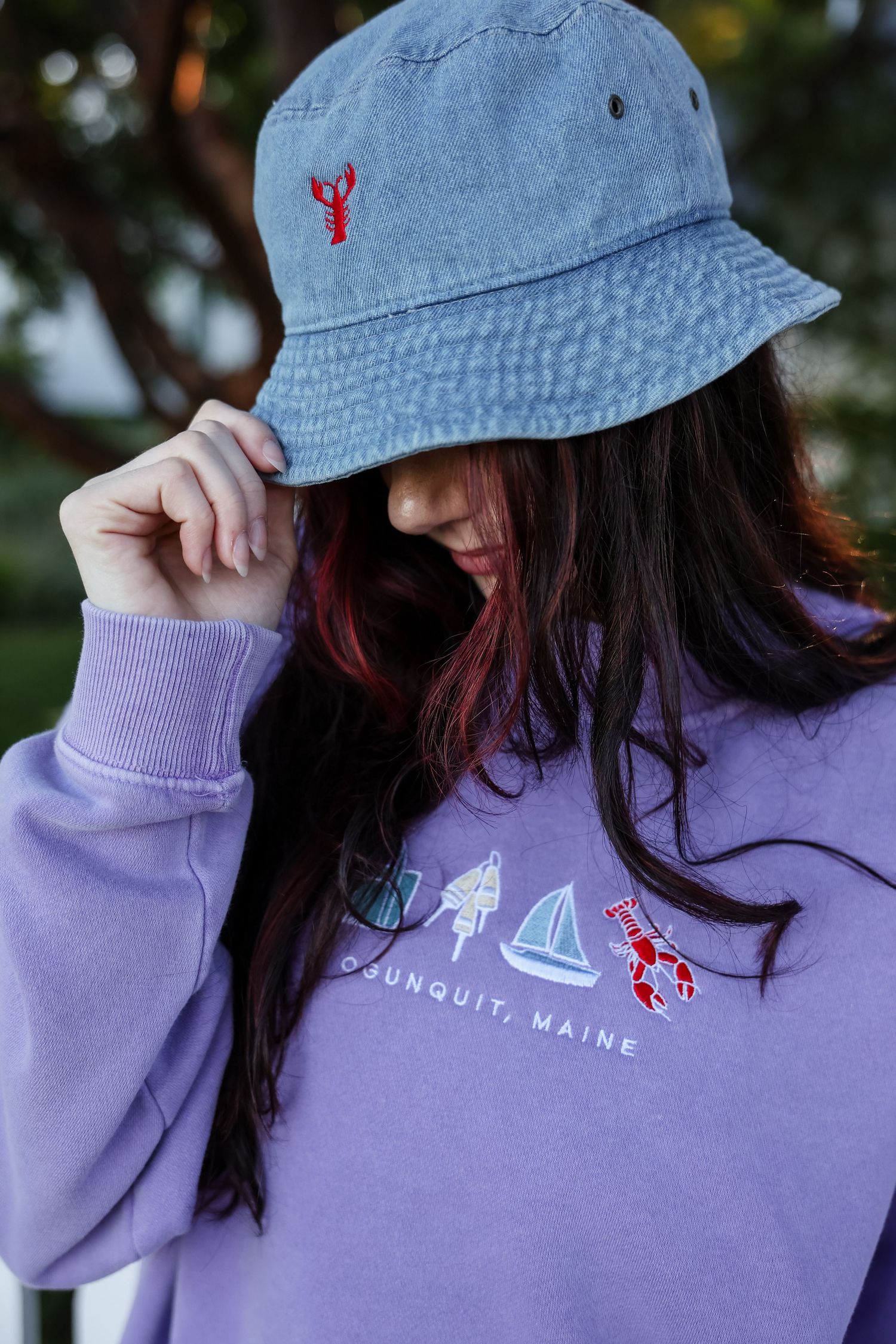 Woman in a denim bucket hat with an embroidered red lobster and a lavender Ogunquit, Maine sweatshirt with sailboat and lobster designs.