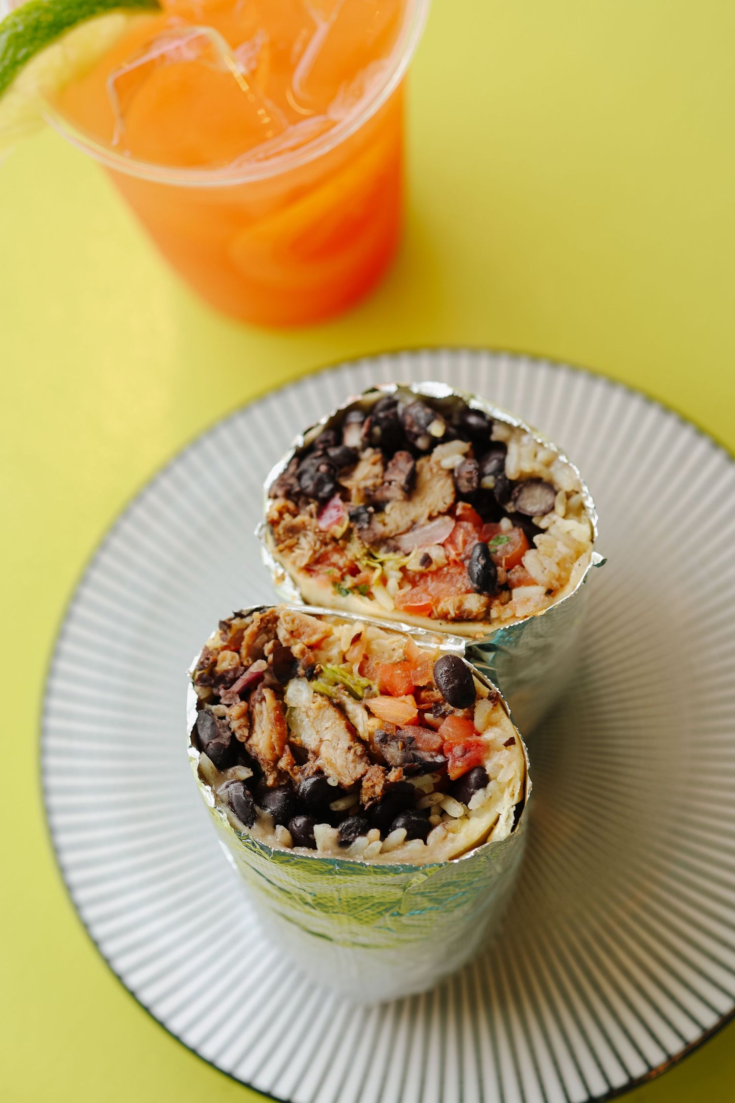 A burrito wrap filled with black beans, rice, and vegetables served on a white plate with orange juice on yellow background.