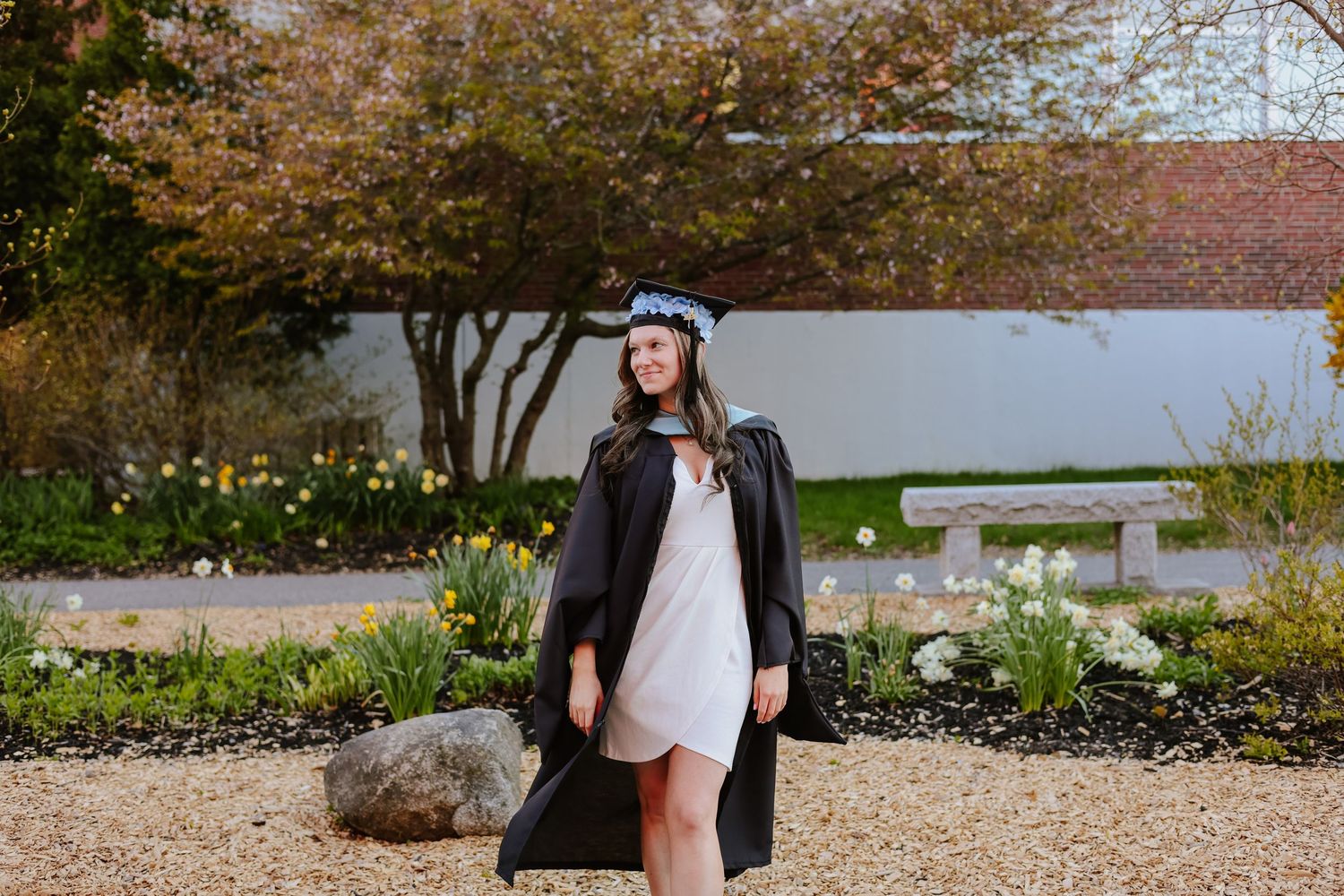 A smiling graduate in black cap and gown poses outdoors among spring flowers and trees.