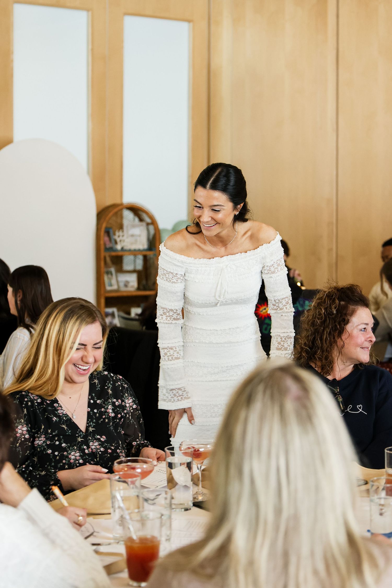 Woman in white off-shoulder dress smiling at guests during a bridal brunch celebration.