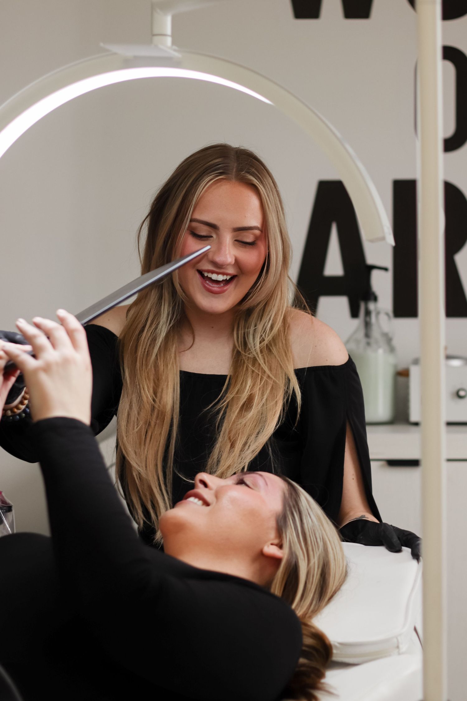 Eye brow artist laughing while working on a client's eyebrows at a modern salon with white decor.