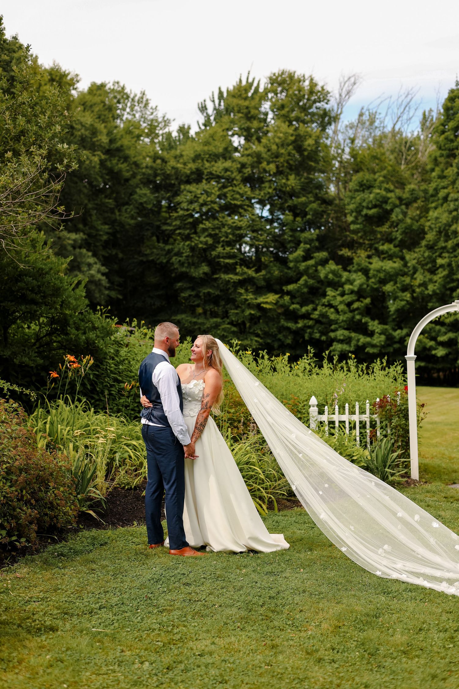 Bride and groom share a romantic moment in a lush garden, her long veil flowing gracefully beside a white arbor.