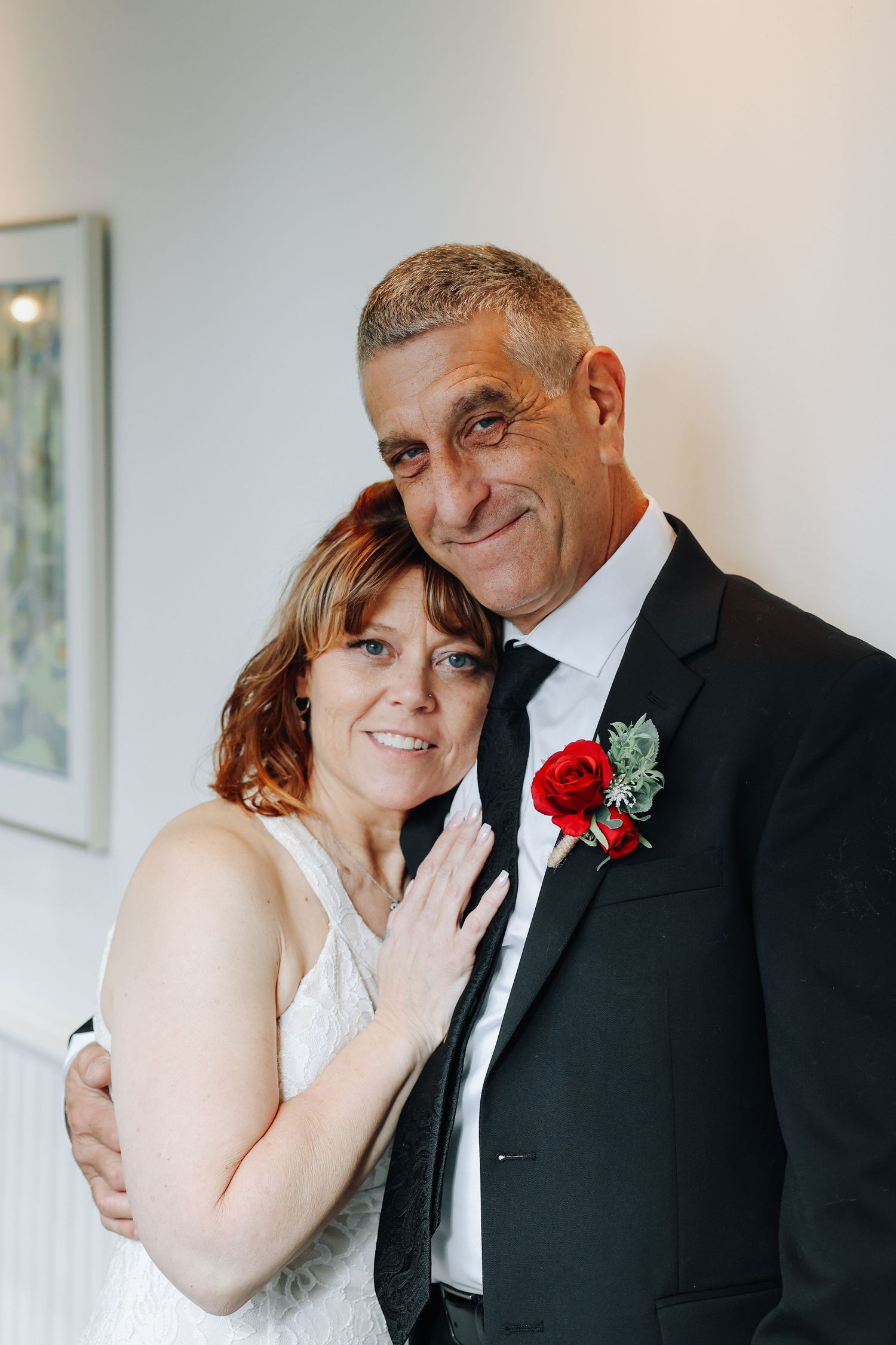 A bride in white and groom in black suit with red boutonniere pose together for an intimate wedding portrait.
