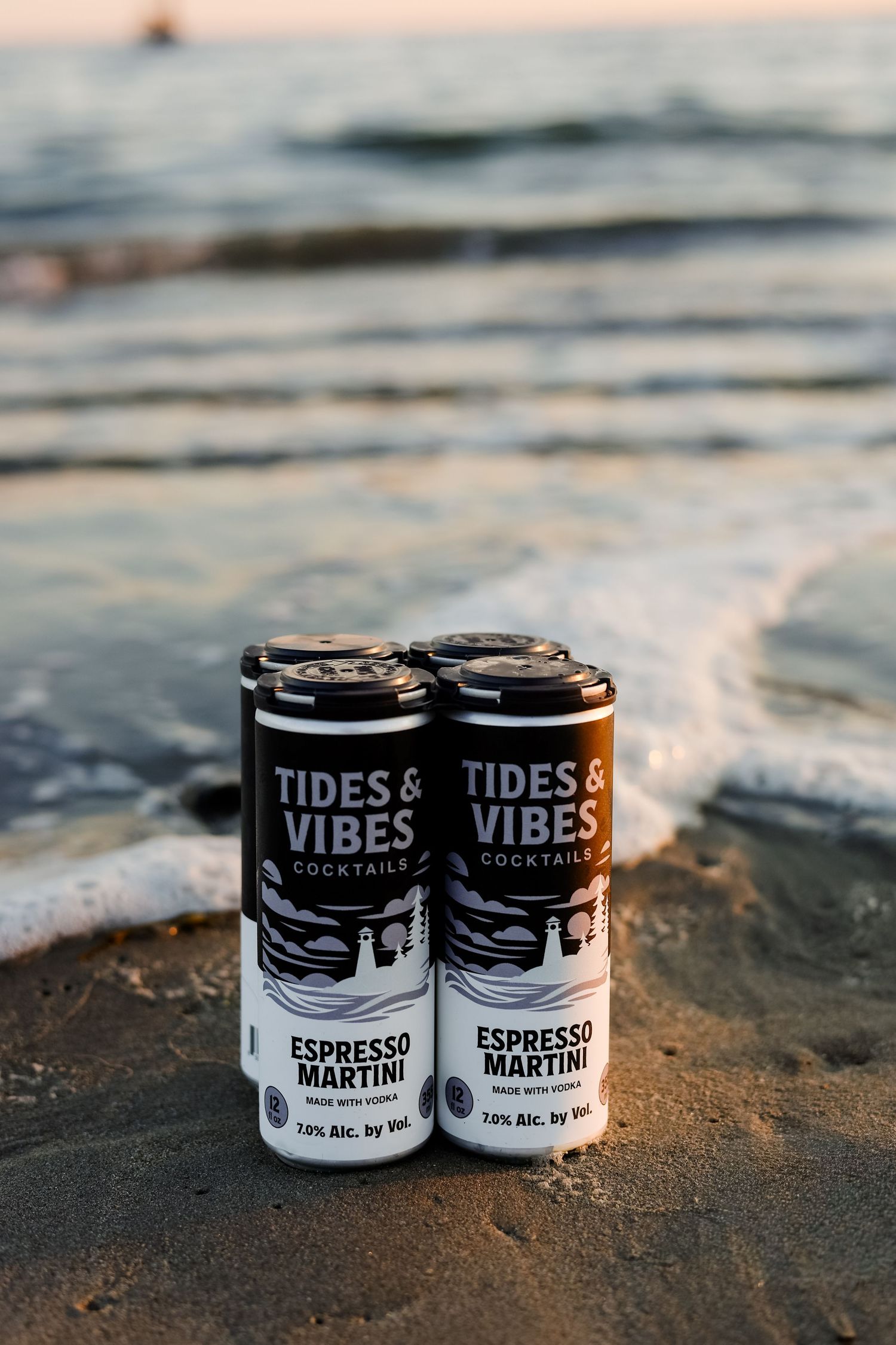 Tides & Vibes Espresso Martini canned cocktails sitting in ocean sand with waves in the background.