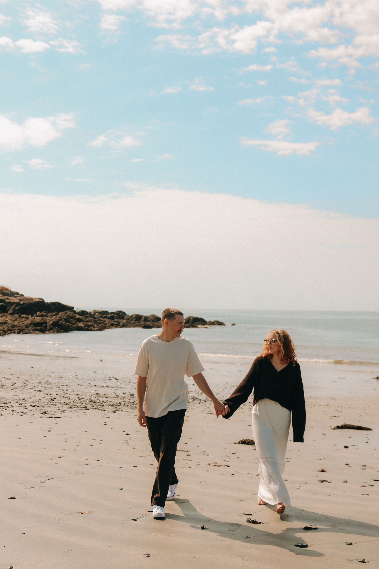 Couple walking hand in hand along a sunny beach with blue skies overhead.