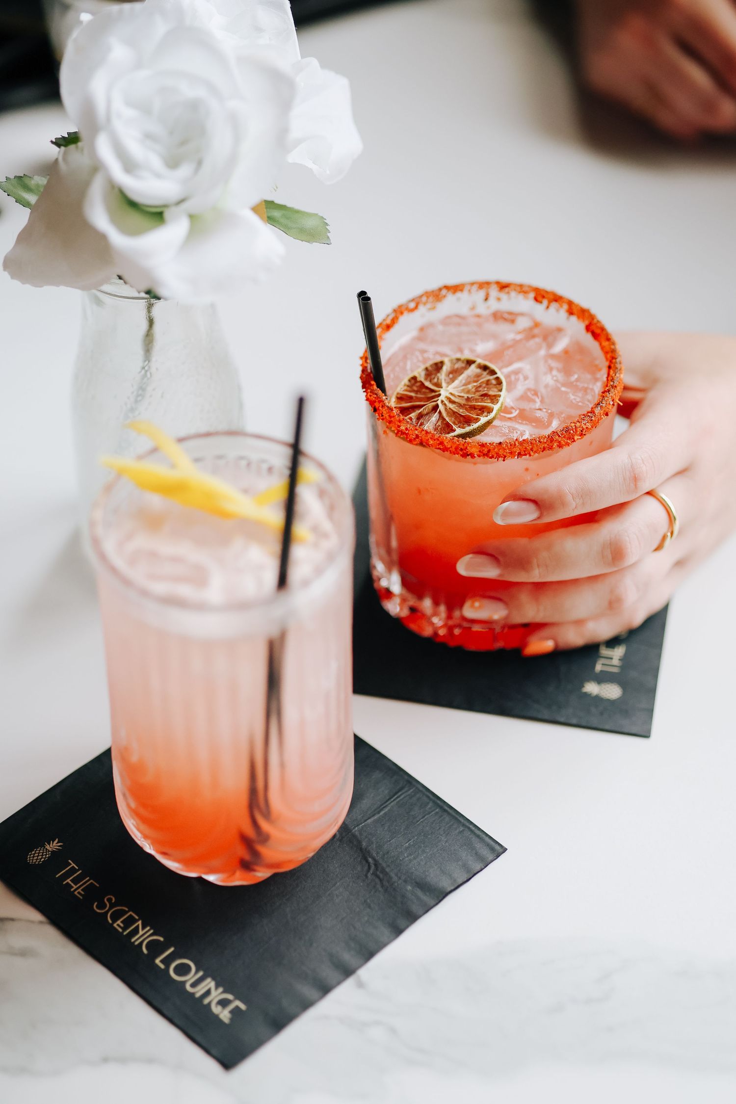 Two craft cocktails with citrus garnish and salt rim served with white rose decoration.