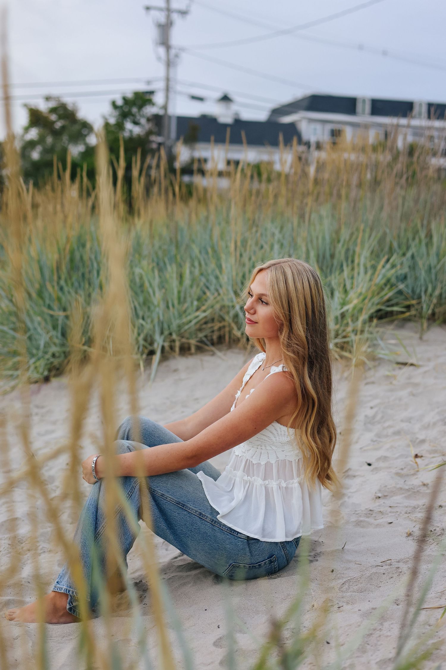 Young woman with long blonde hair sits on sandy beach in white top and jeans, surrounded by tall dune grass.