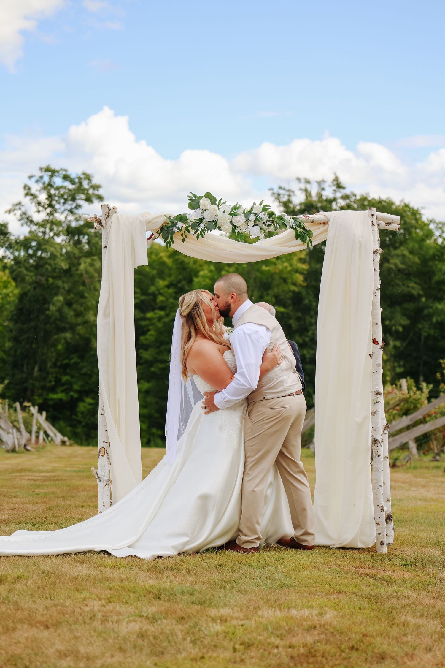 Wedding ceremony site with white draped arch and floral decorations in sunny field.
