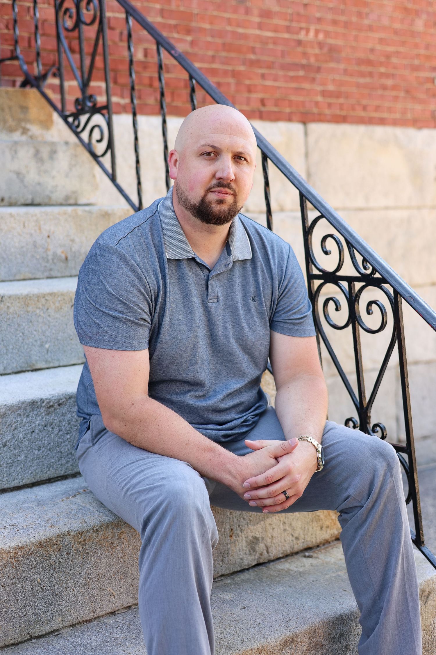 Bald bearded man in a gray polo and pants sits with clasped hands on granite steps beside an ornate wrought iron railing and brick wall.