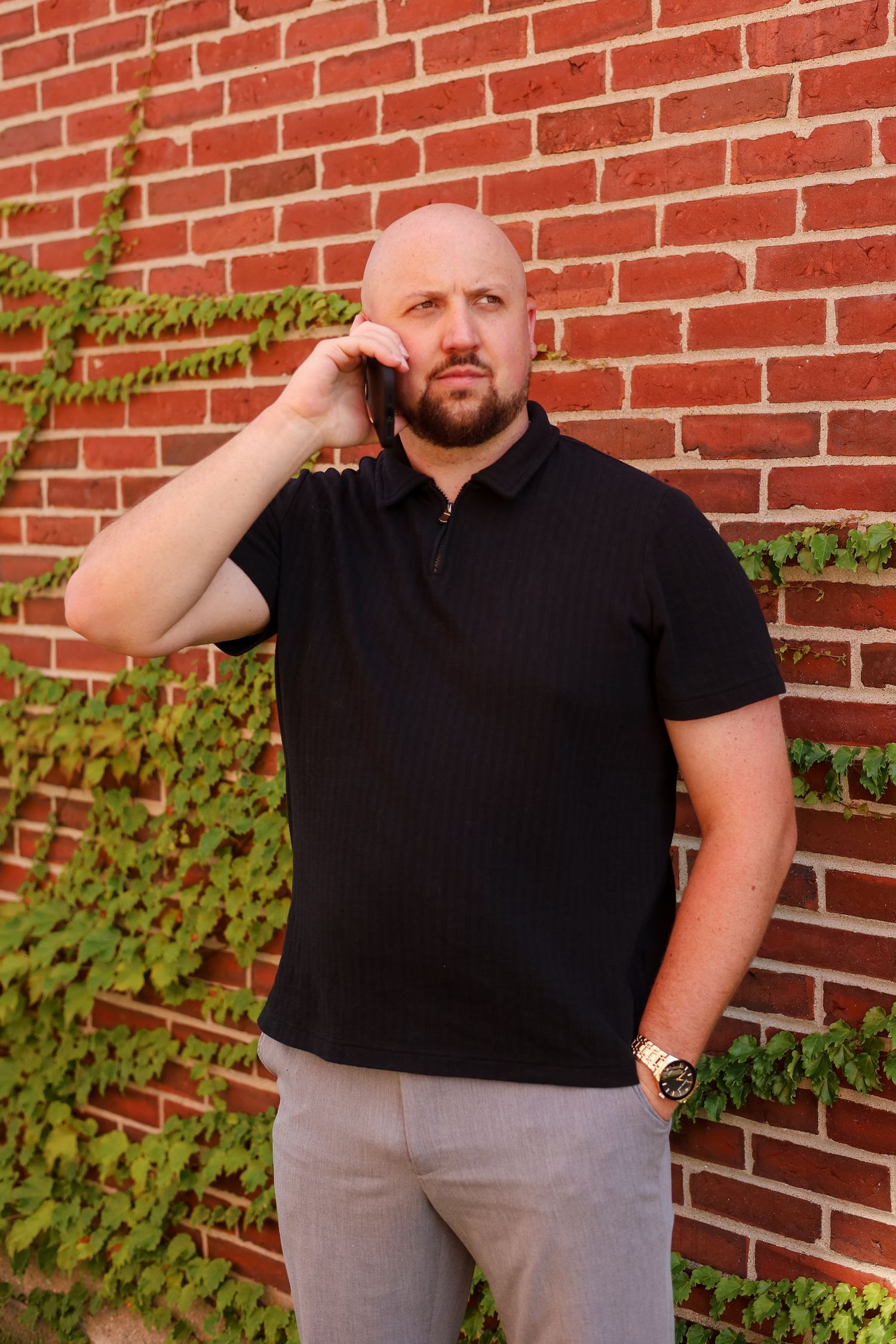 Bald bearded man in black polo shirt talking on smartphone against red brick wall with ivy.