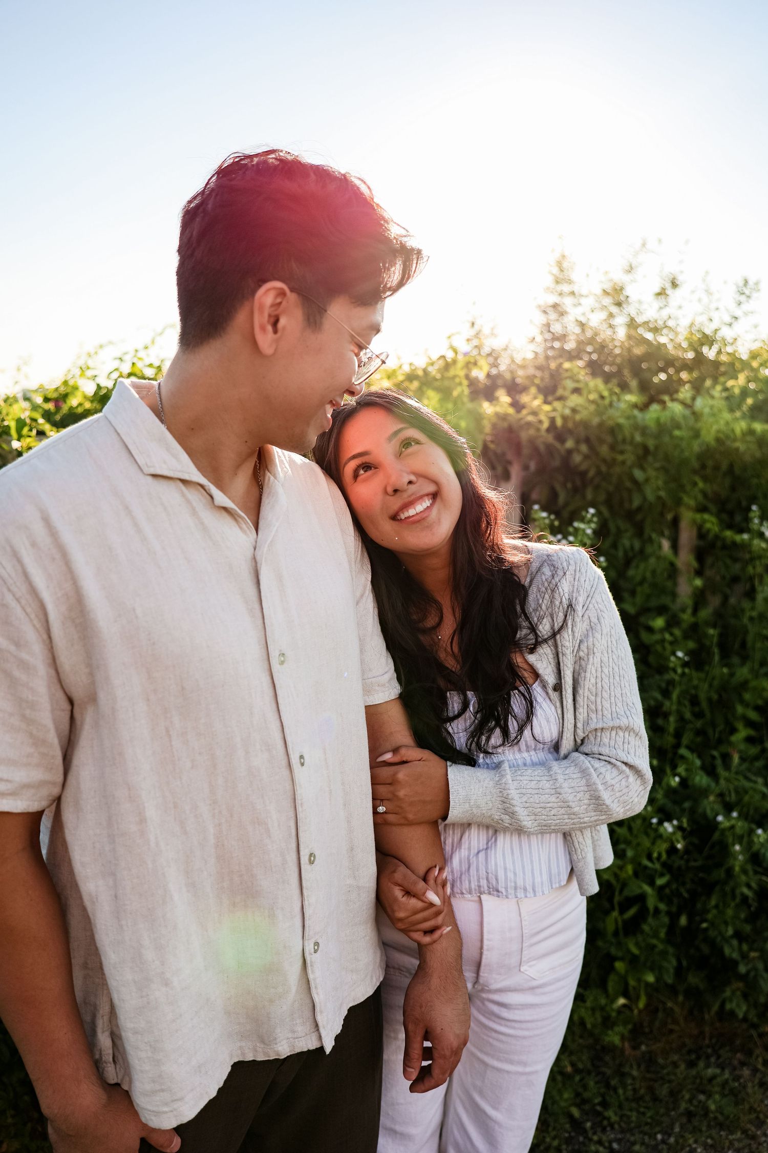 A happy couple embraces outdoors in golden sunlight, smiling at each other near lush green hedges.