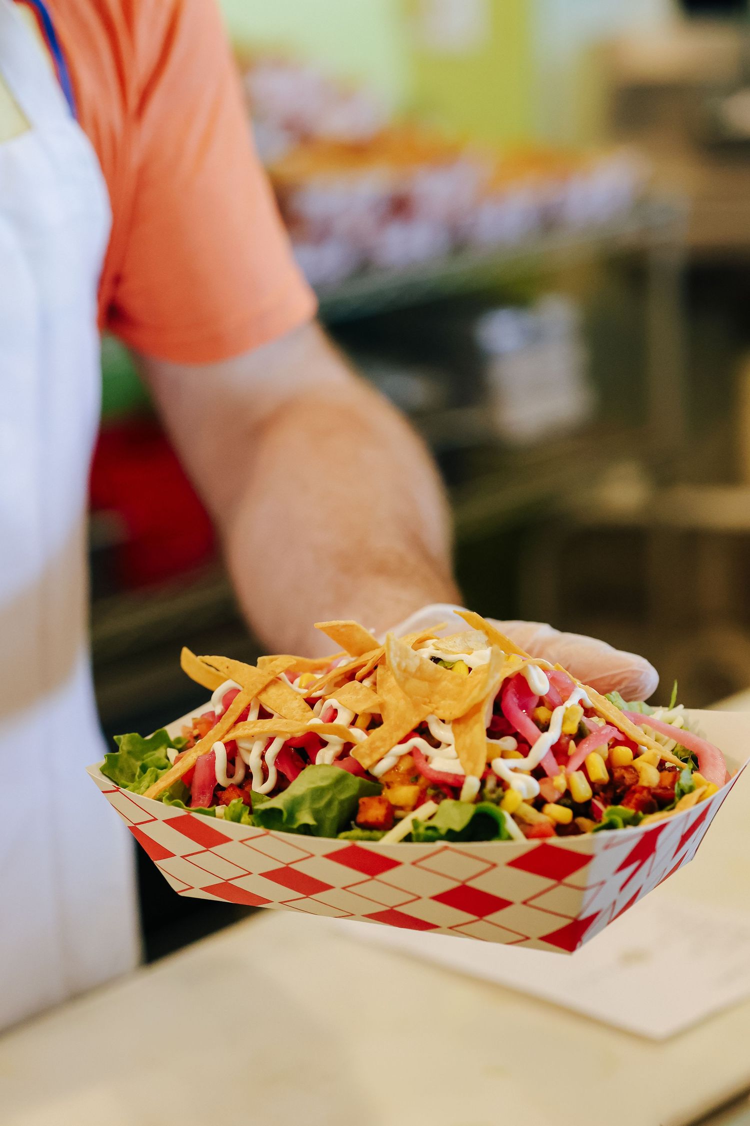 A colorful taco salad served in a red and white striped paper basket with shredded cheese, lettuce and fresh toppings.