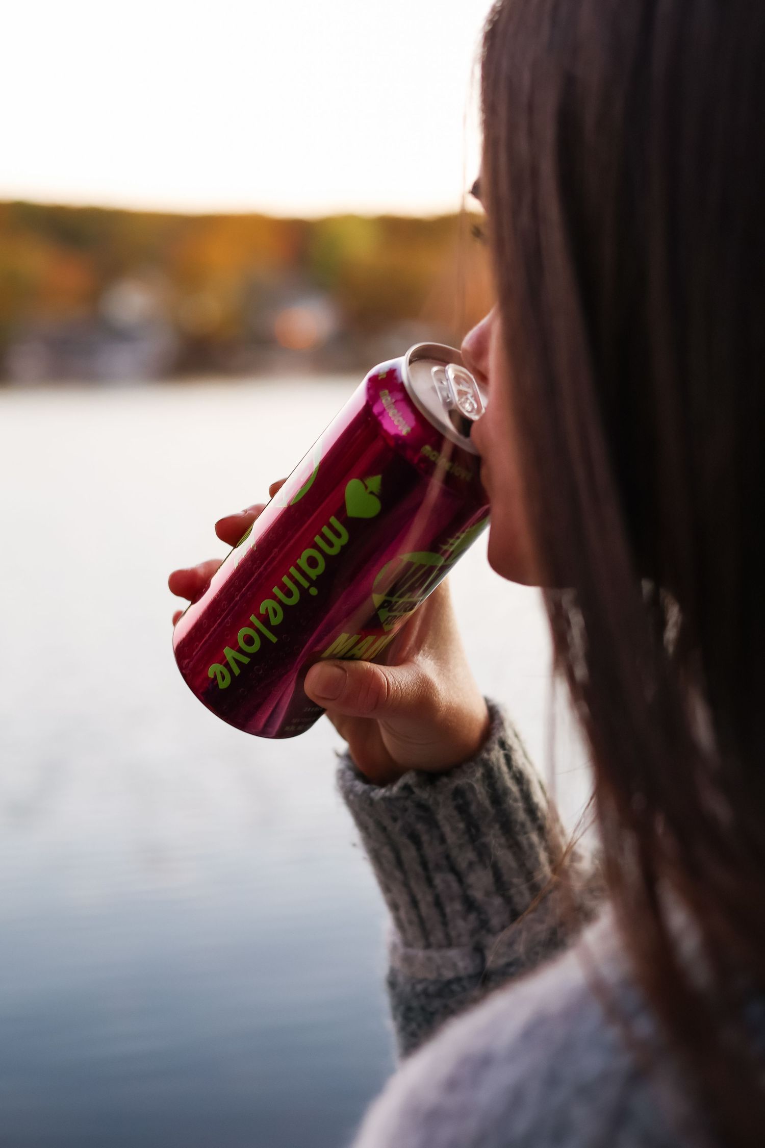 Woman in a gray knit sweater sips from a purple Maine Love can beside a calm lake with blurred autumn foliage.