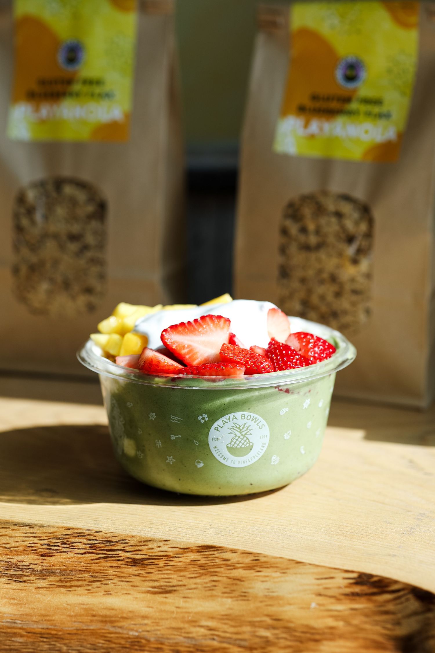 Green matcha smoothie bowl topped with fresh strawberries, mango, and cream, served in a plastic cup on a wooden board.