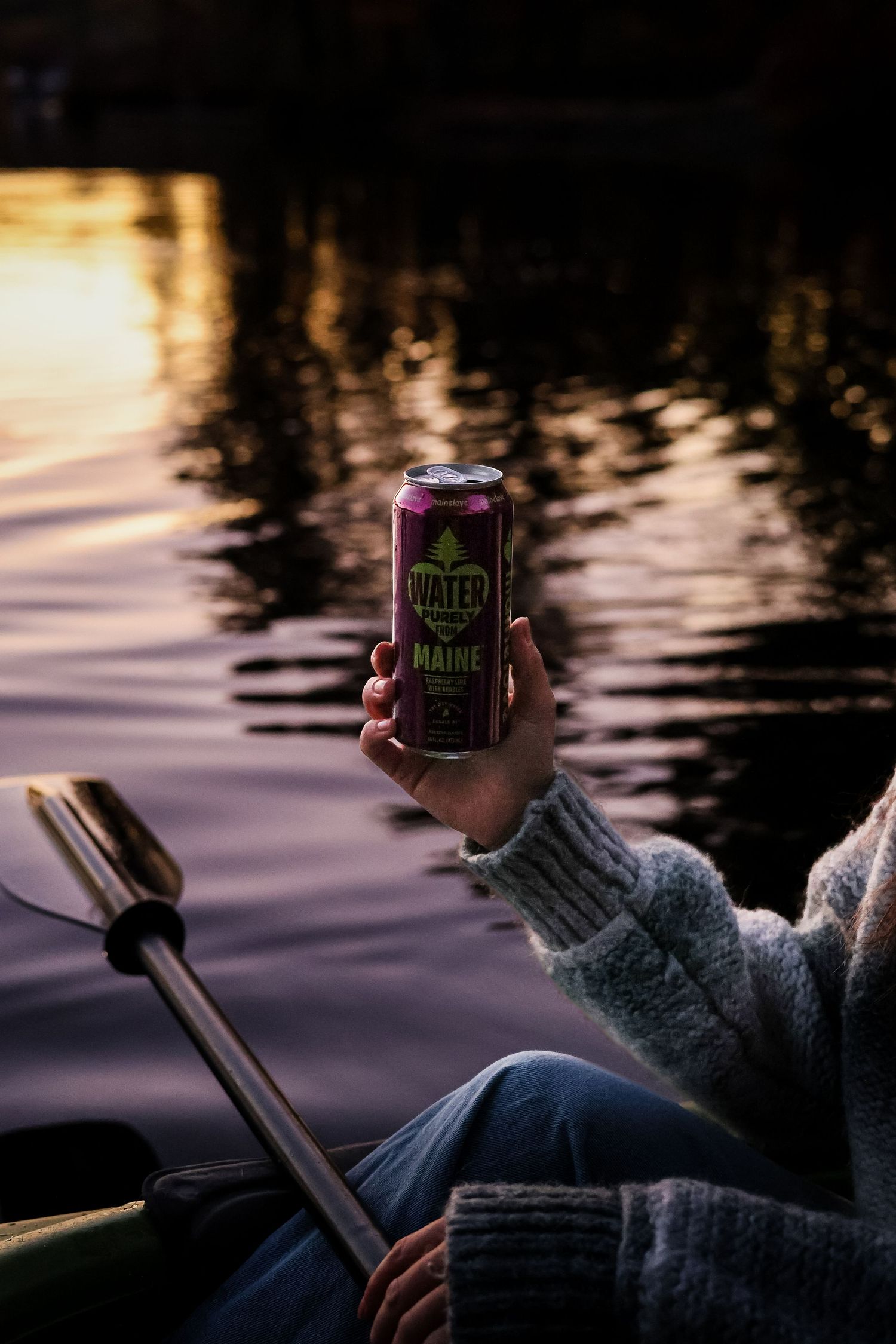 Person in a cozy gray sweater holds a Maine water can above a calm lake at sunset while seated in a kayak with a paddle.