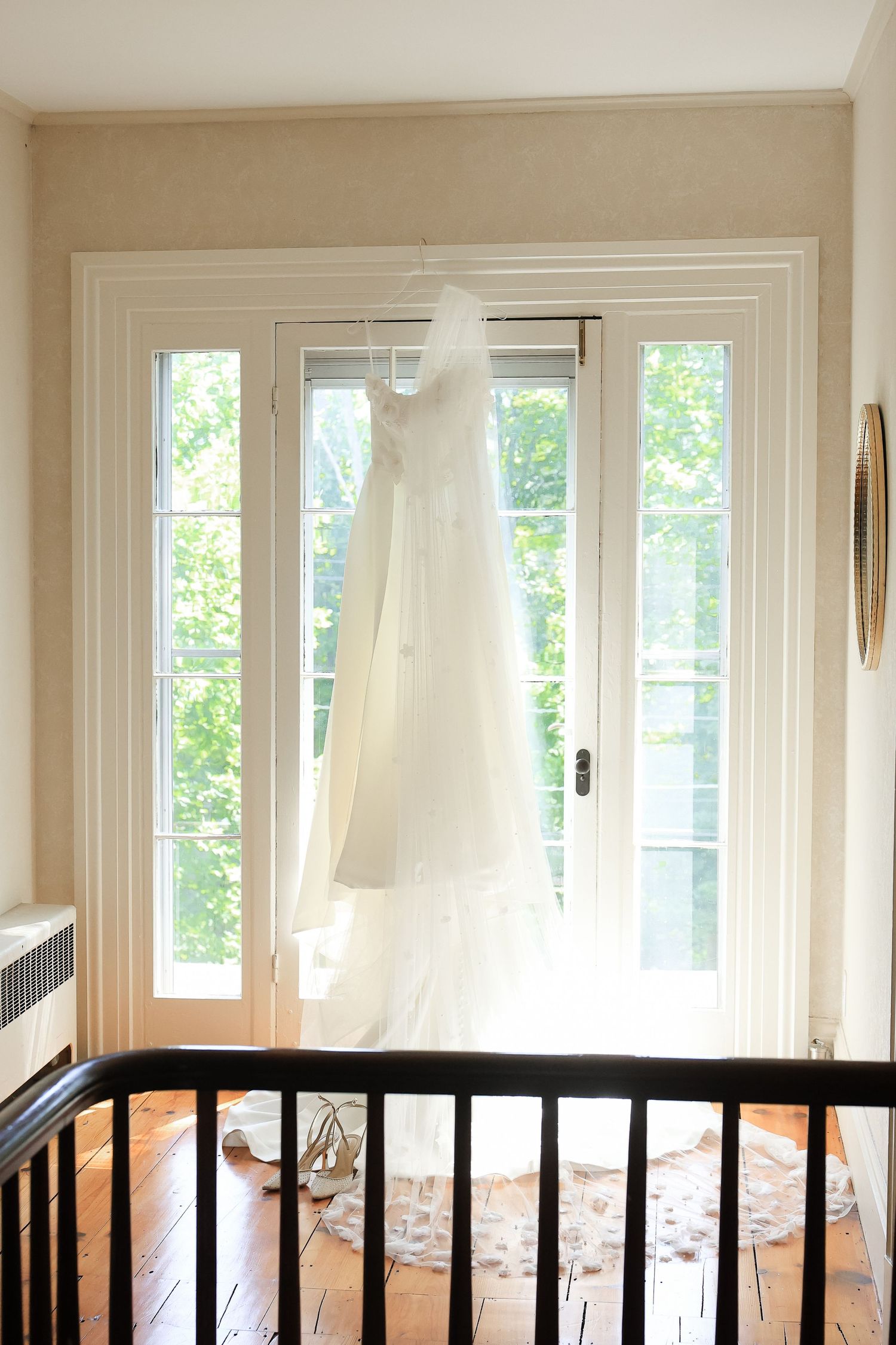 A white wedding dress hanging on French doors, with natural light streaming through, wooden floors, and a staircase railing in foreground.