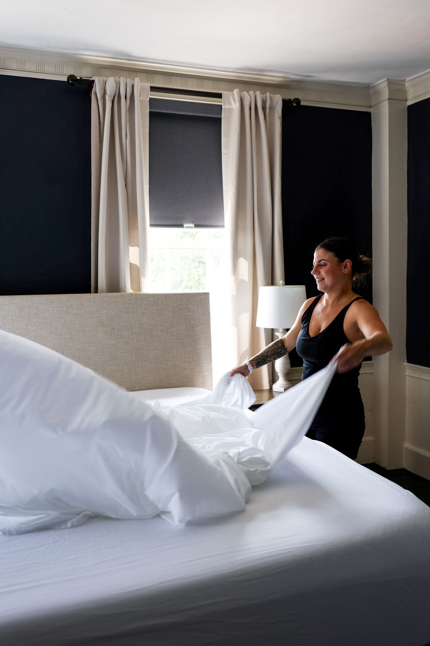 A person making a bed with white linens in a room with dark walls and white curtains, shown in a sequence of images.