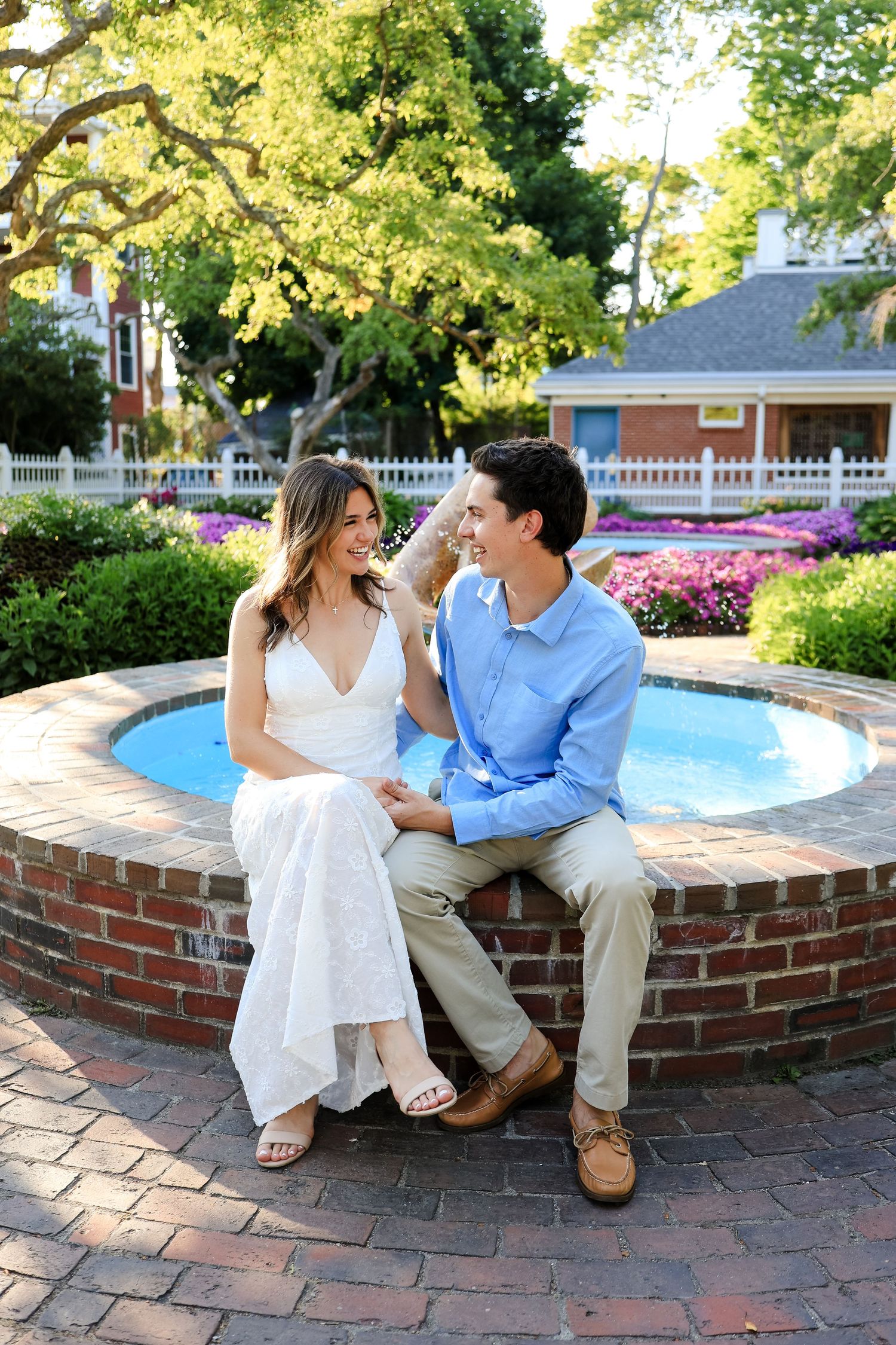 A couple sits together on a brick fountain, smiling at each other in a garden setting.