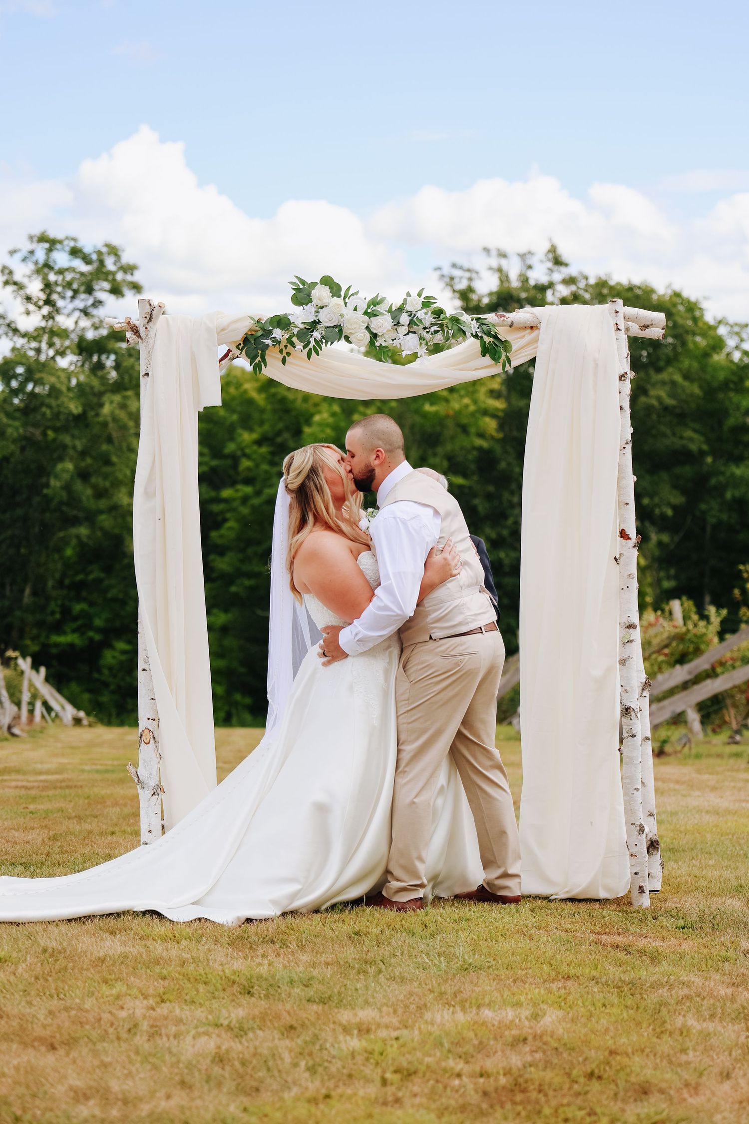 Bride and groom share a kiss under a white draped wedding arch adorned with greenery and flowers outdoors.