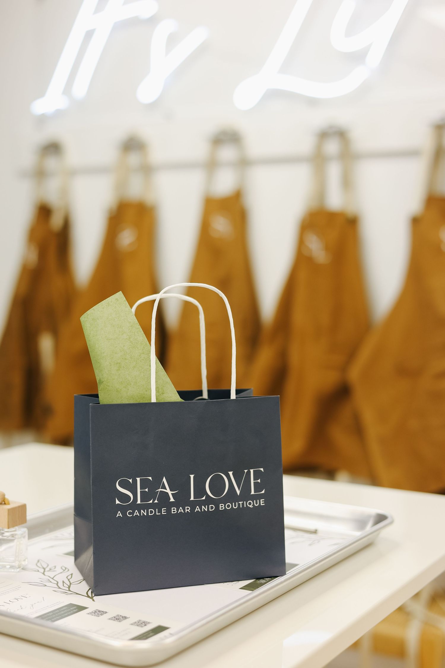 A navy Sea Love Candle Bar and Boutique gift bag sits on a tray with brown aprons hanging in the background.