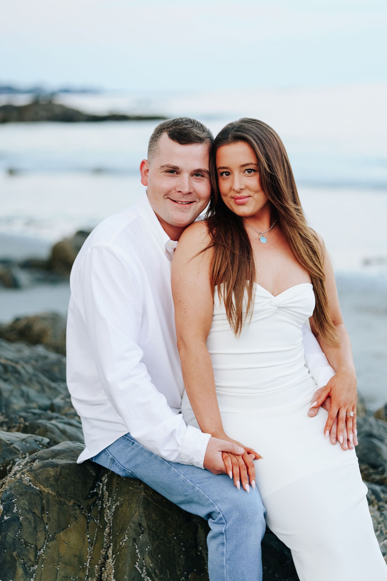 Couple in white outfits posing on rocky beach, woman in white dress leaning on man in white shirt and jeans.