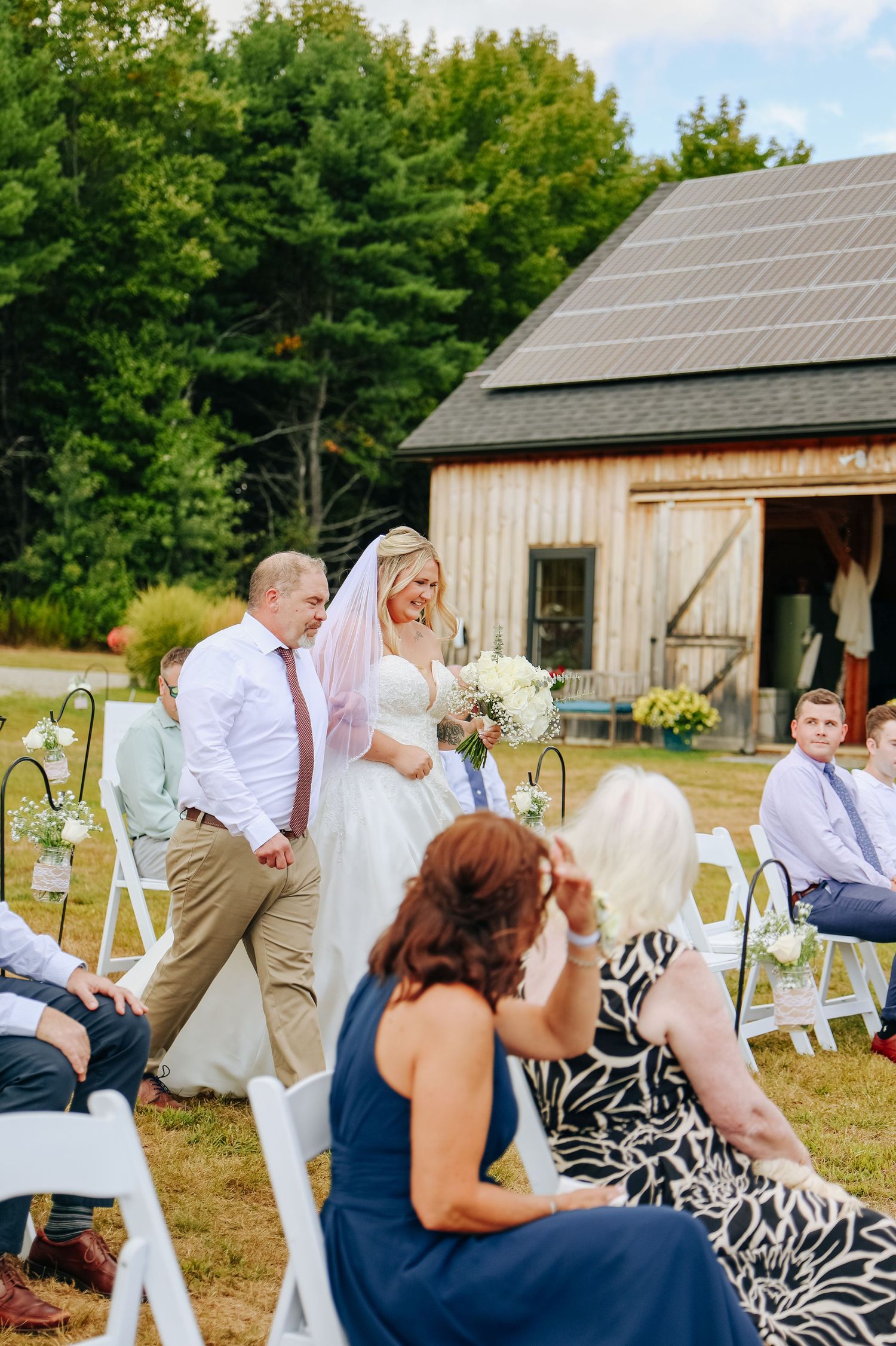 Outdoor wedding ceremony setup with white chairs and rustic barn in background.