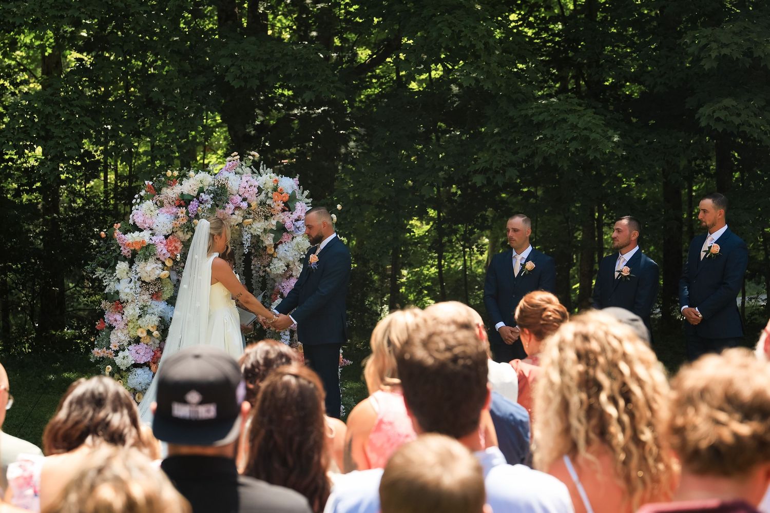 Outdoor wedding ceremony with bride and groom at floral arch, groomsmen standing nearby, guests seated in sunlit forest setting.