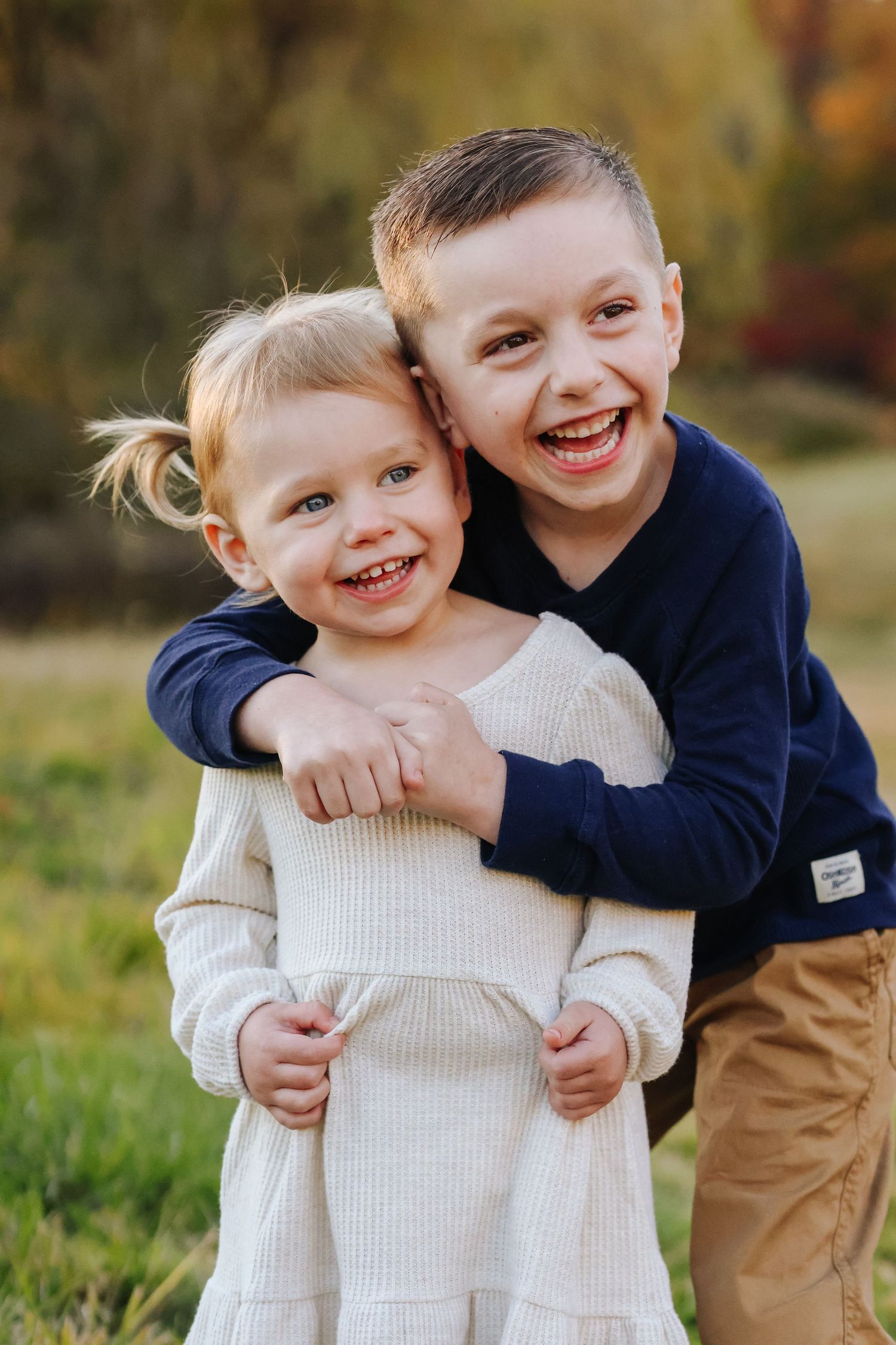 Portrait of two children embrace and laugh together while wearing cozy sweaters outdoors.