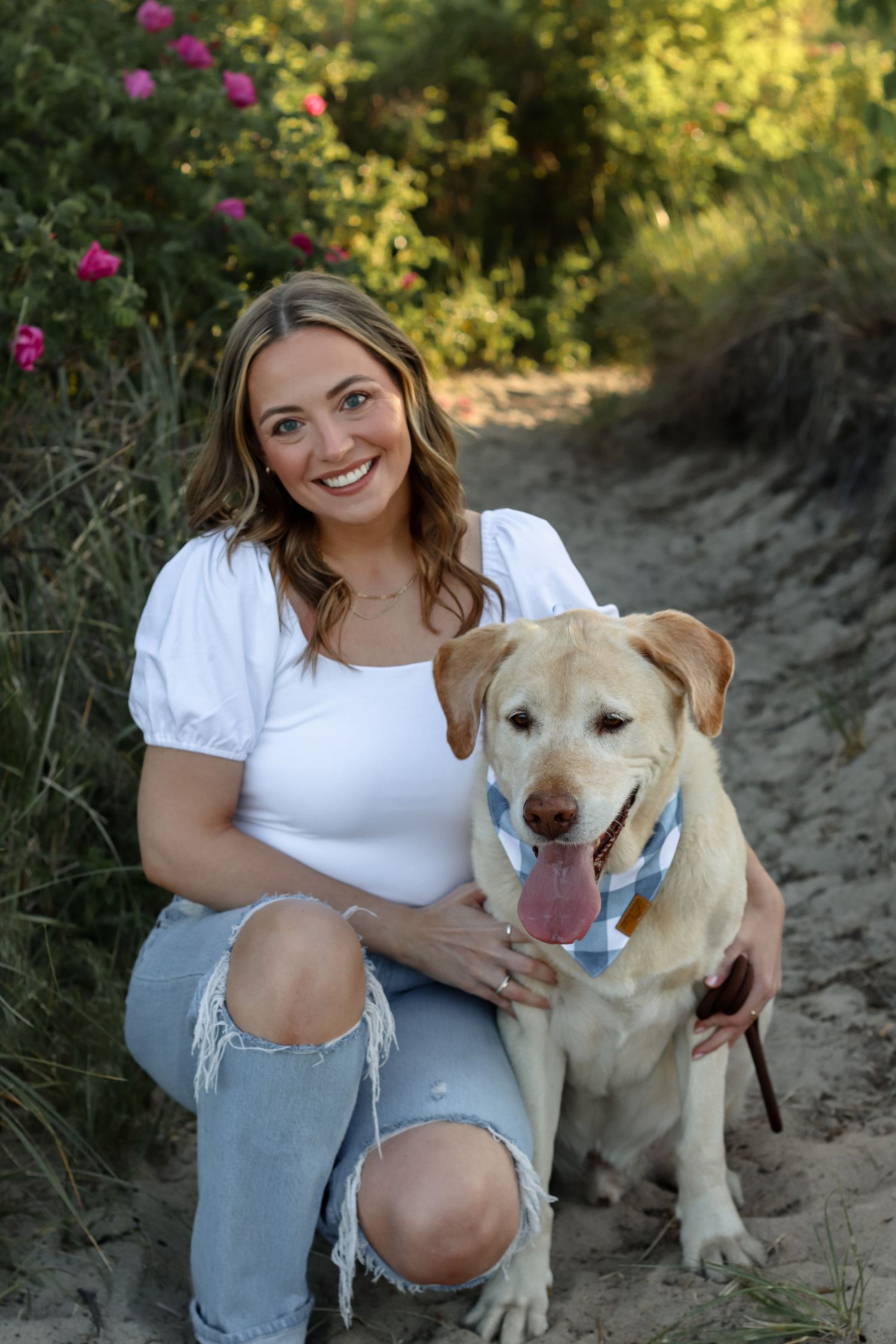 A person in casual attire sits with a friendly yellow Labrador dog among blooming flowers.