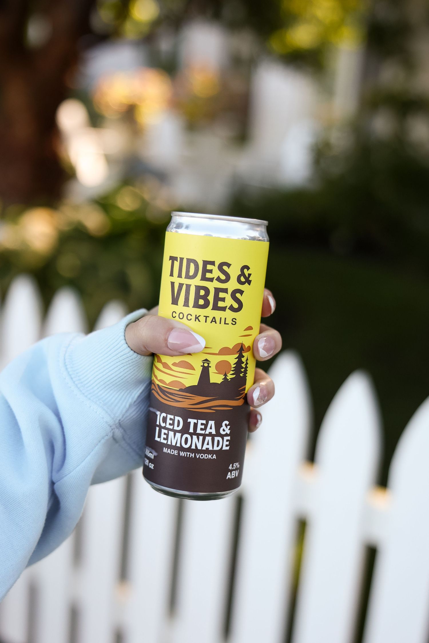 Hand holding a Tides & Vibes iced tea and lemonade vodka cocktail can in front of a white picket fence.