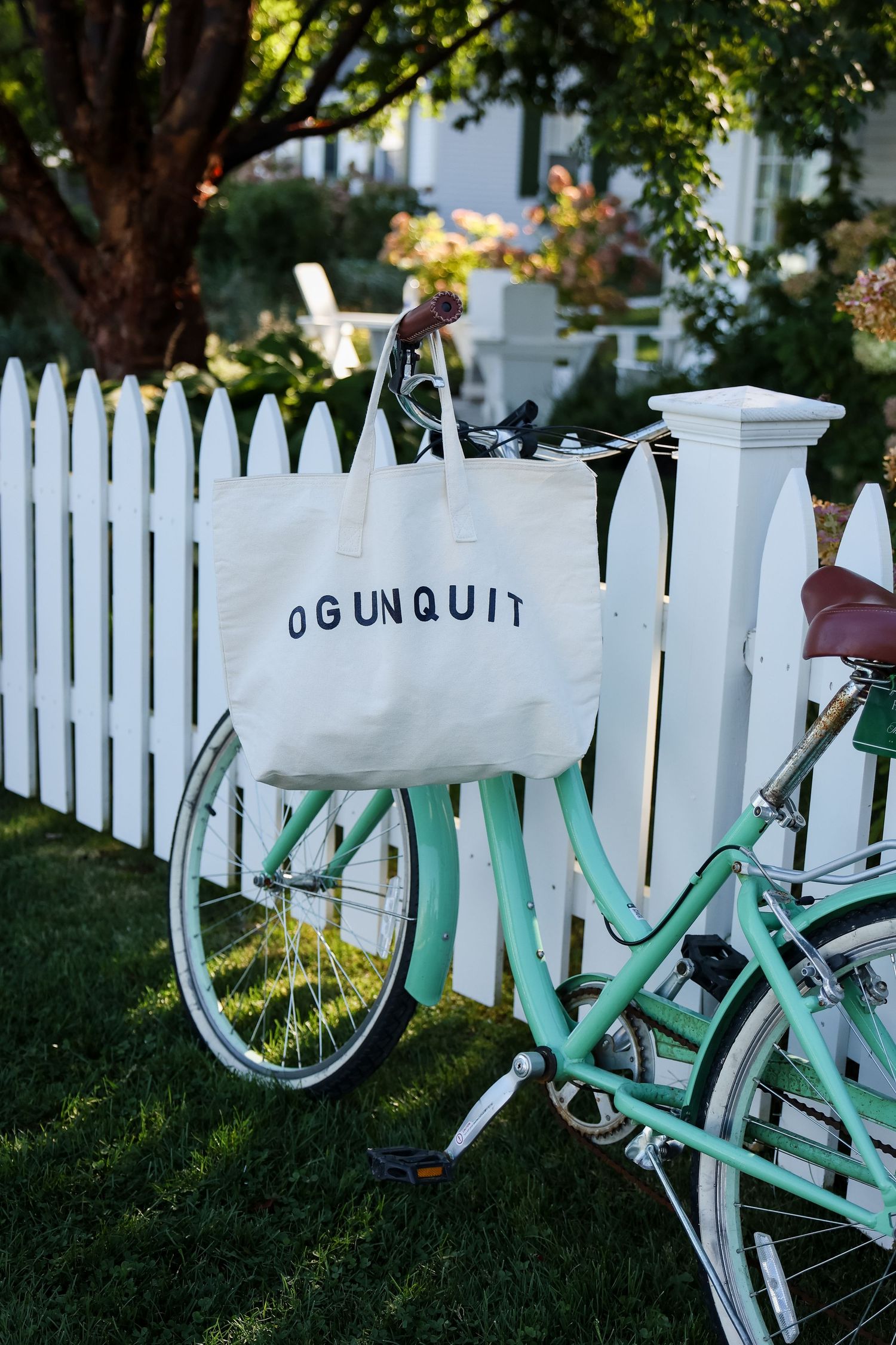 Mint green vintage bicycle with an Ogunquit canvas tote bag hanging from the handlebars, parked against a white picket fence.